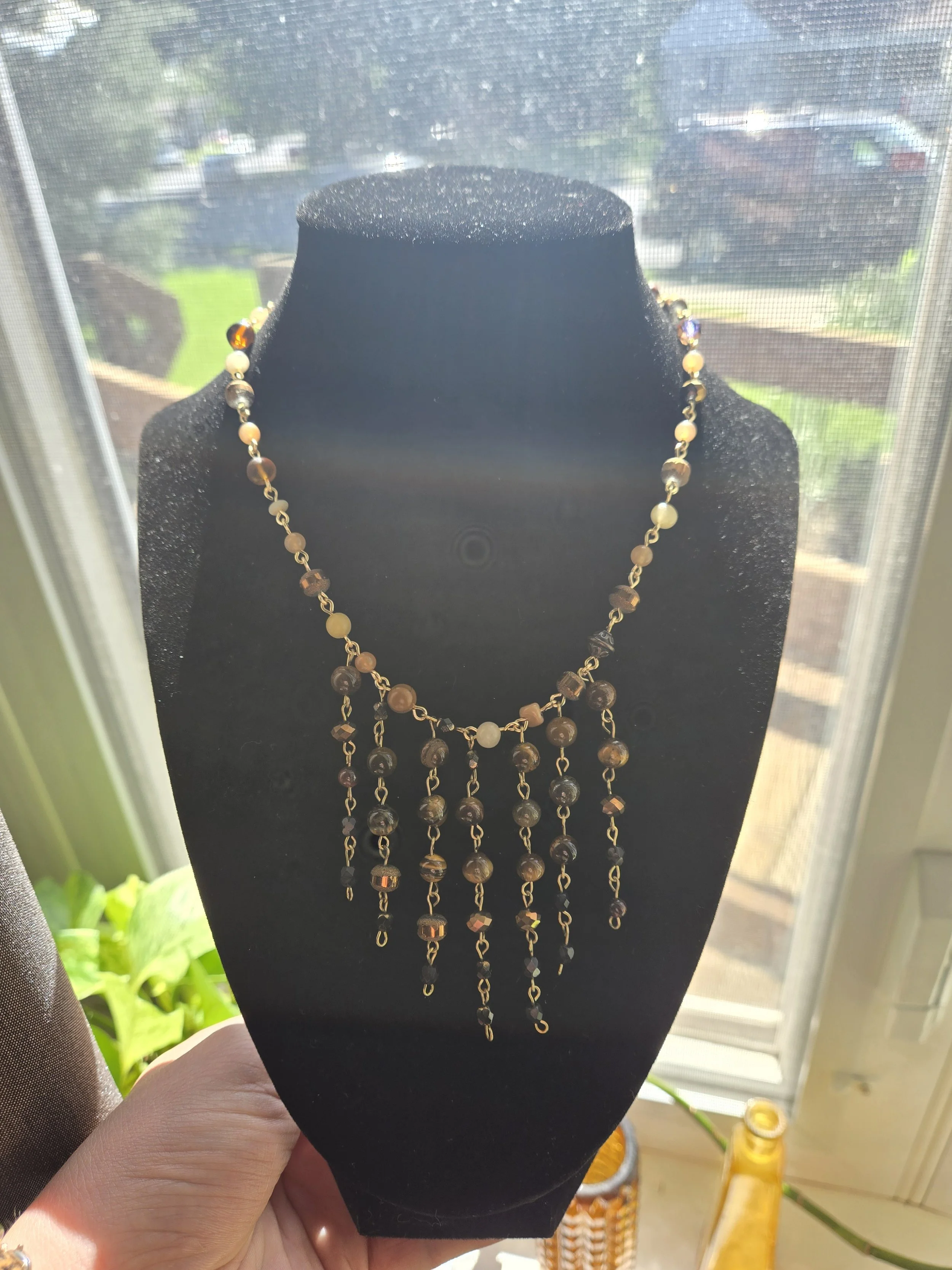 Necklace with multiple strands of beads and gold-colored chains displayed on a black bust, with a window and outdoor scenery in the background.
