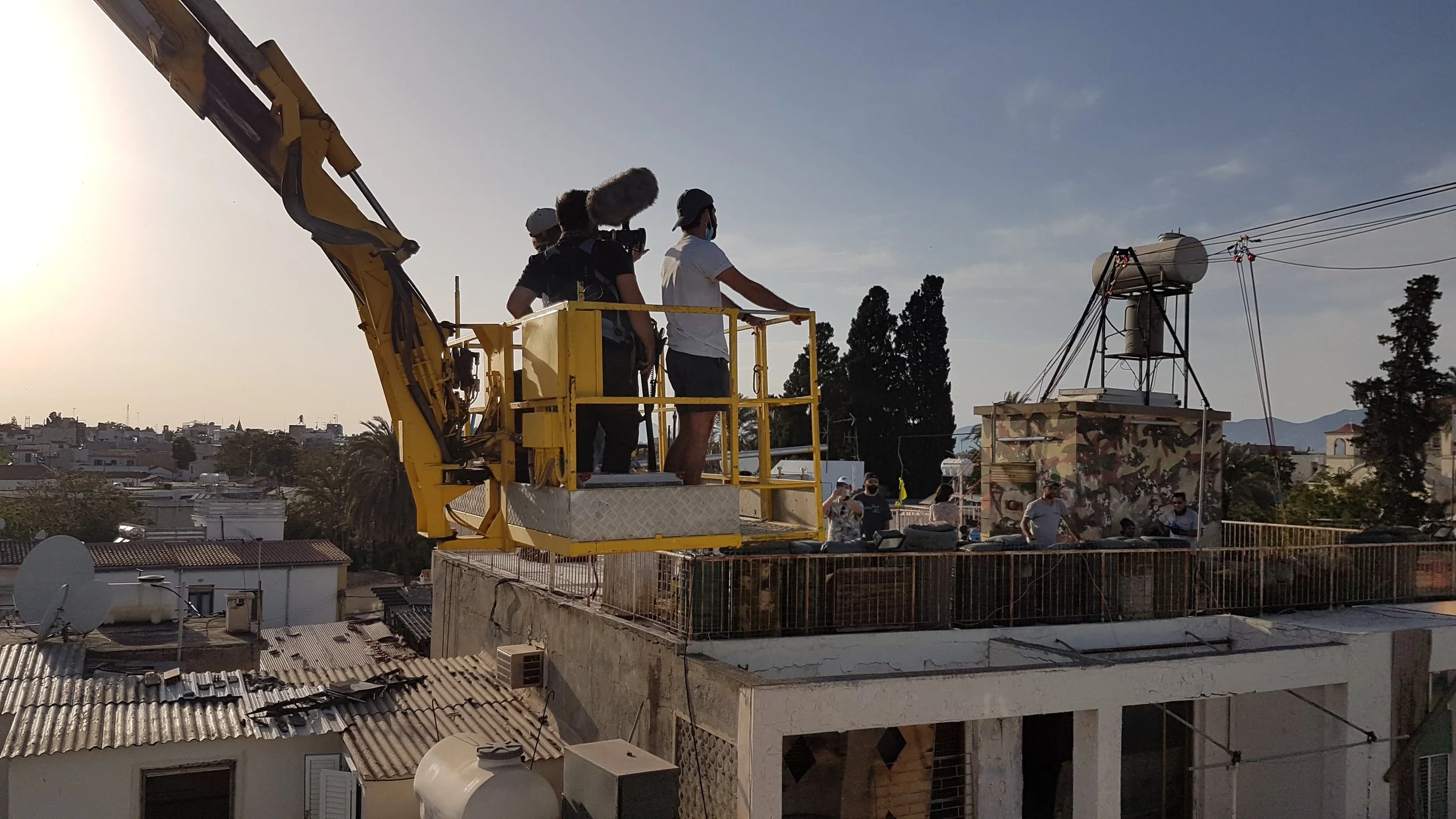A camera crew filming on a rooftop with a yellow crane, with other crew members and onlookers nearby, under a clear sky.