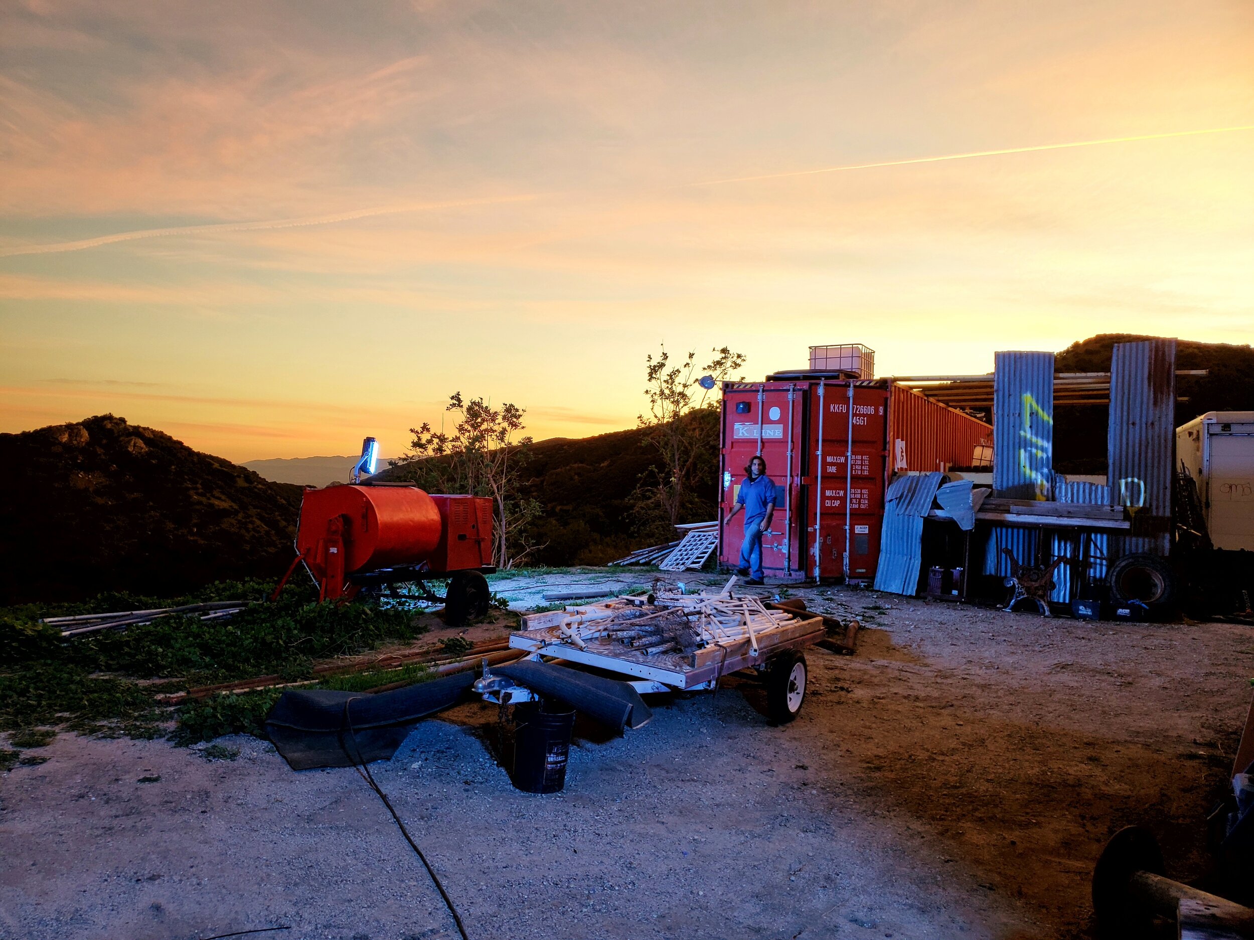 Sunset over a hillside with makeshift structures, including a red shipping container, a red tank, and a small open shed, surrounded by various objects and equipment, with a person standing near the container.