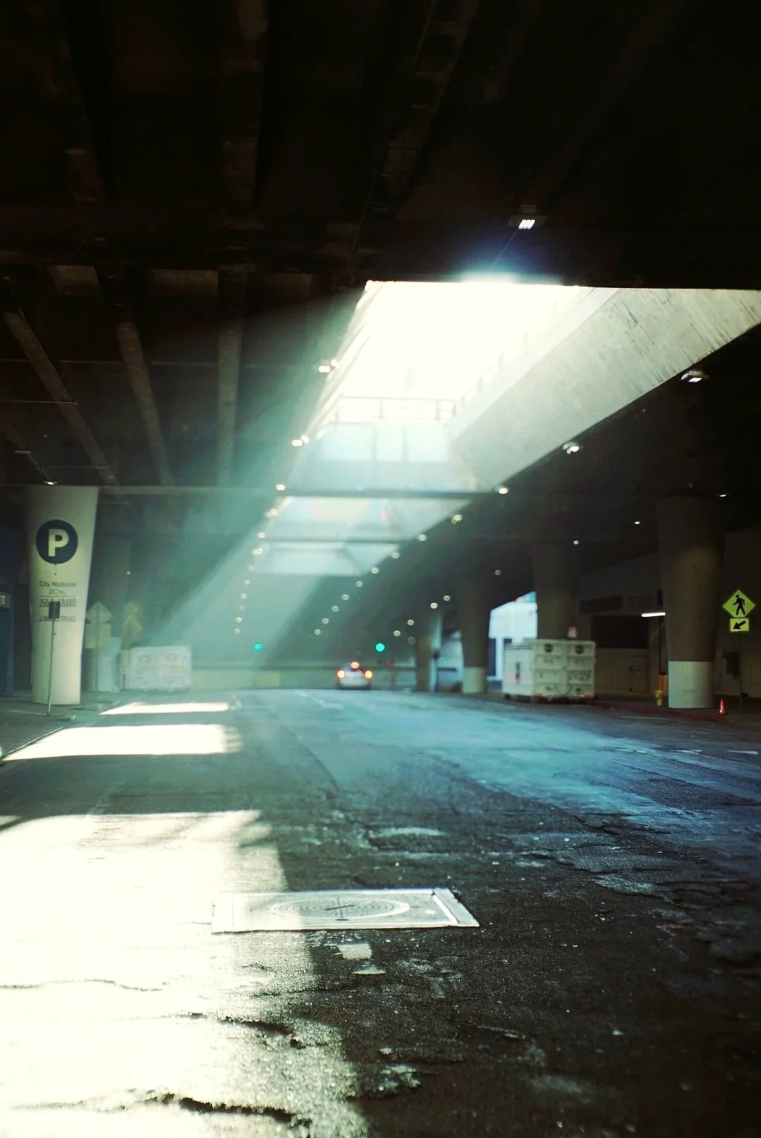 Underpass with sunlight streaming through an opening above, illuminating the street below with a car in the distance and various signs.