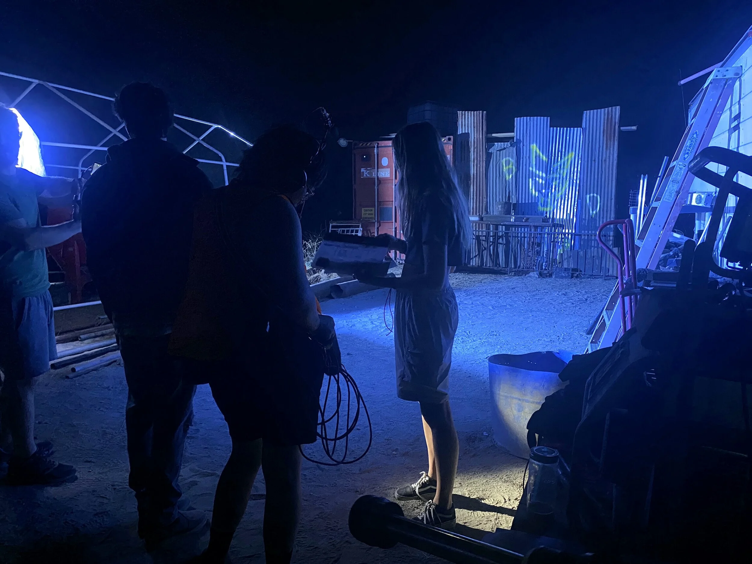 Cast and crew is gathered outdoors at night. This is behind the scenes or during a film shoot, with equipment and lighting visible.
