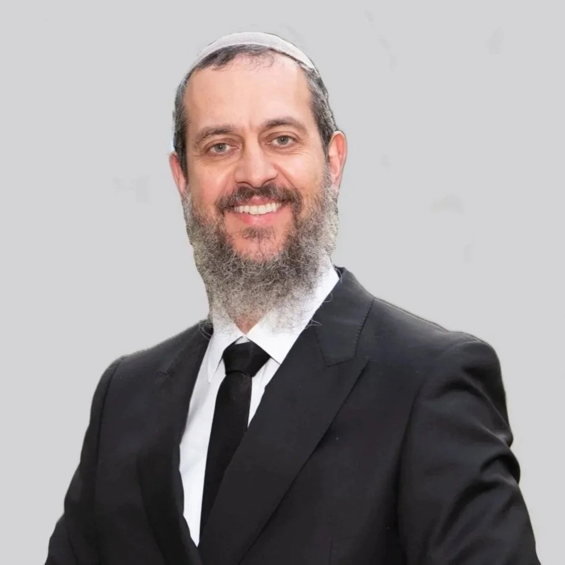 A smiling man wearing a black suit, white shirt, and black tie, with a trimmed beard and wearing a kippah, standing against a light gray background.