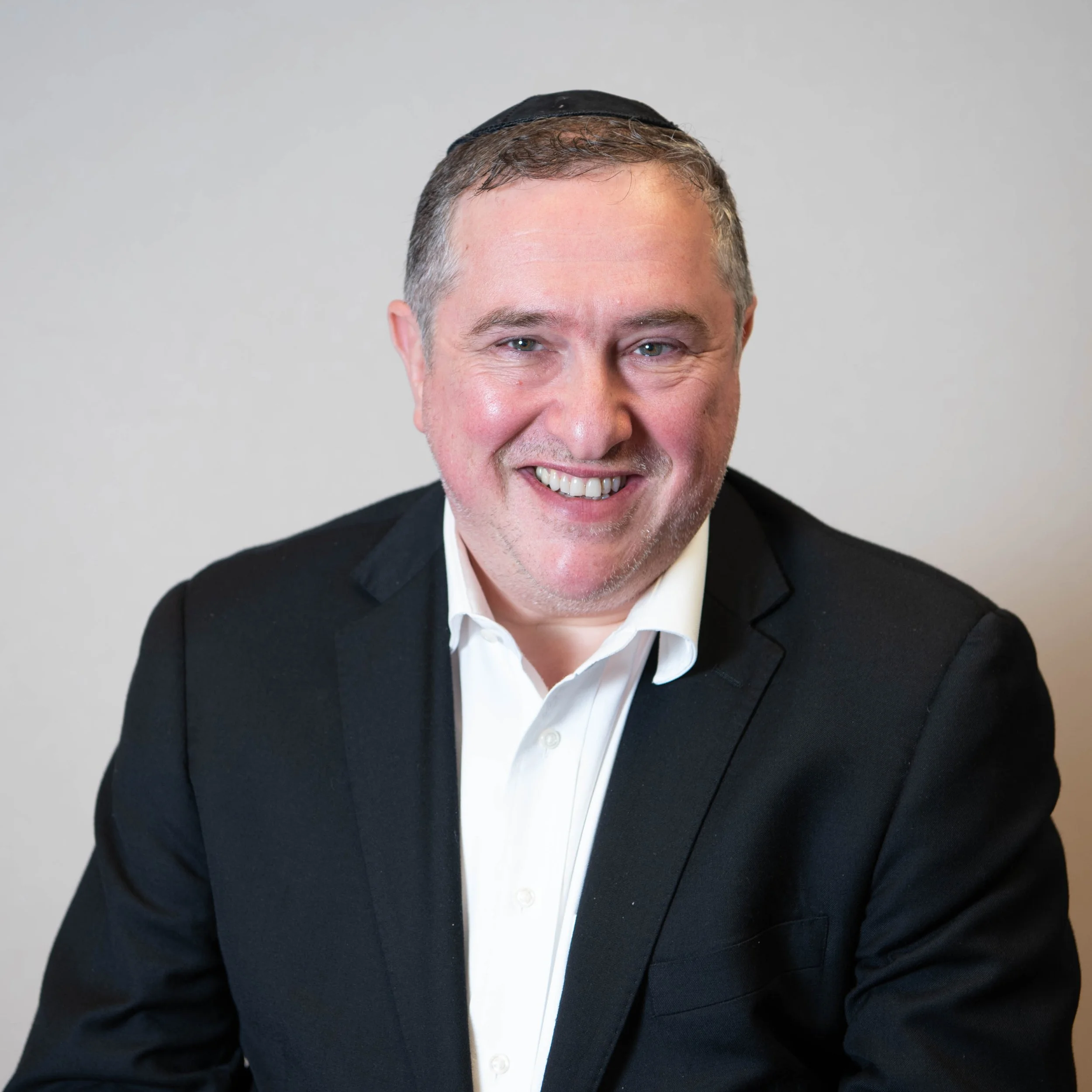 Man with short gray hair, wearing a black suit and white shirt, smiling at the camera, background plain.