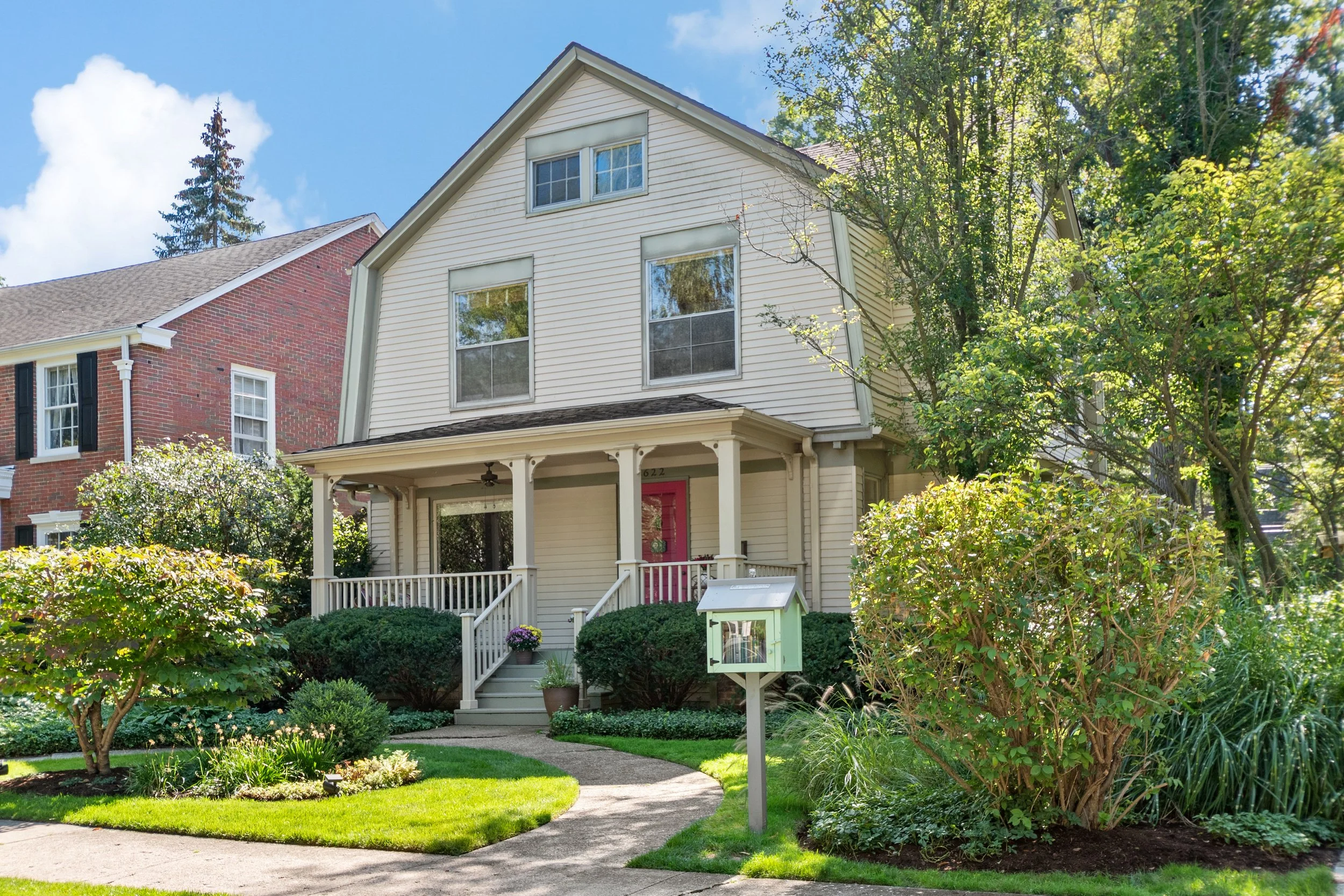 A two-story house with beige siding and a front porch, pink front door, surrounded by a lush green garden and bushes, with a mailbox in the front yard and another house partially visible on the left.