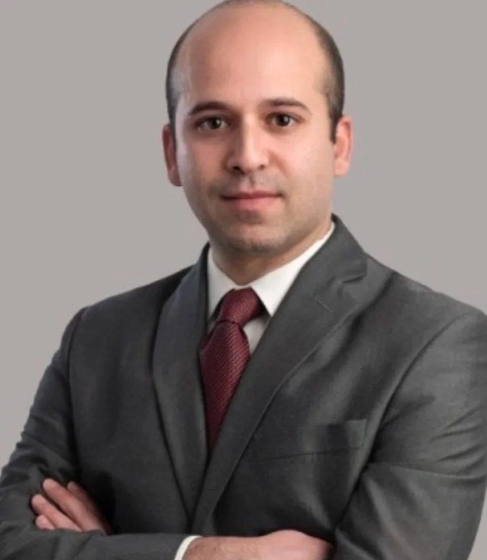 A professional man with light skin, dark eyes, and a shaved face, dressed in a dark gray suit with a white shirt and a red tie, posing with arms crossed against a plain gray background.