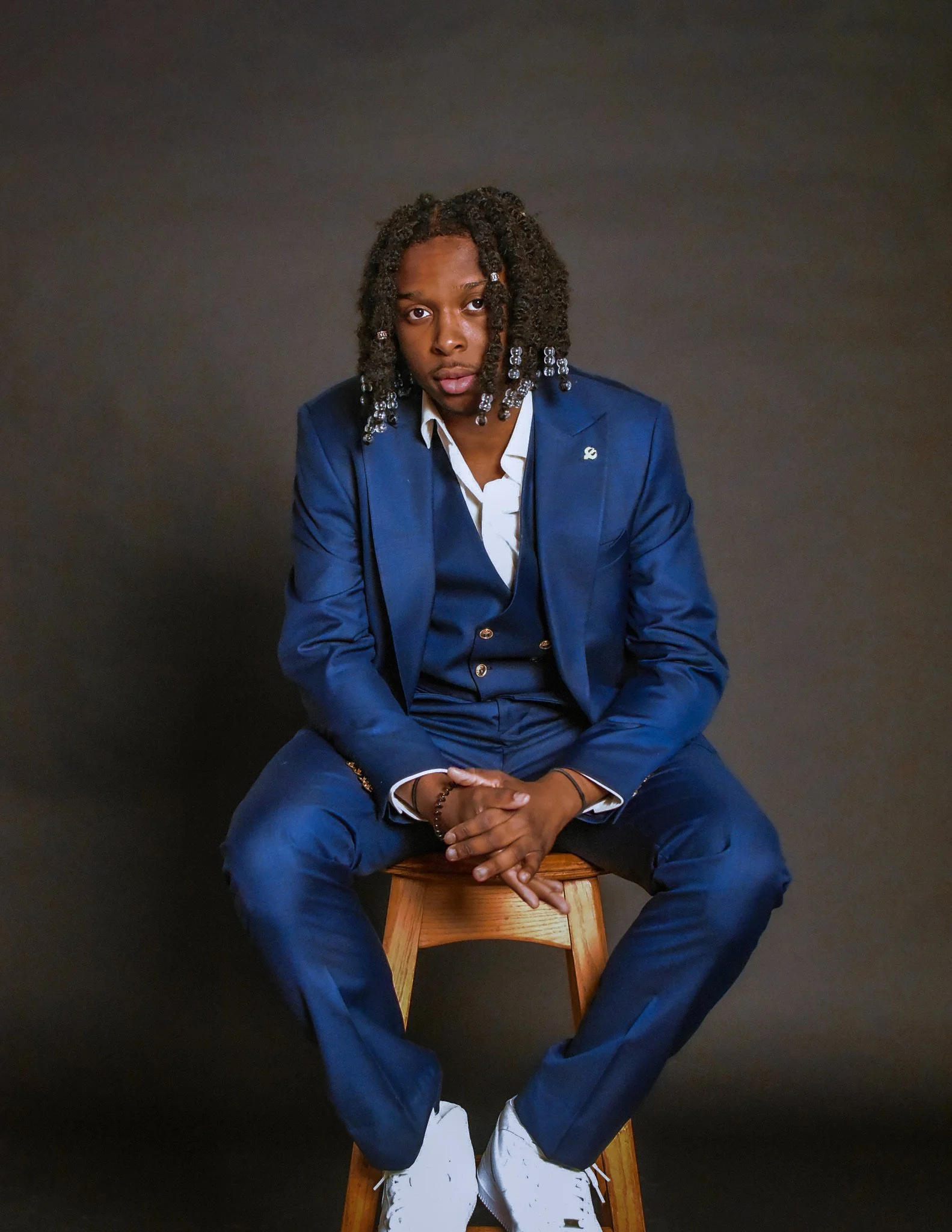 Jeune homme assis sur un tabouret, portant un costume bleu, chaise contre un fond sombre.