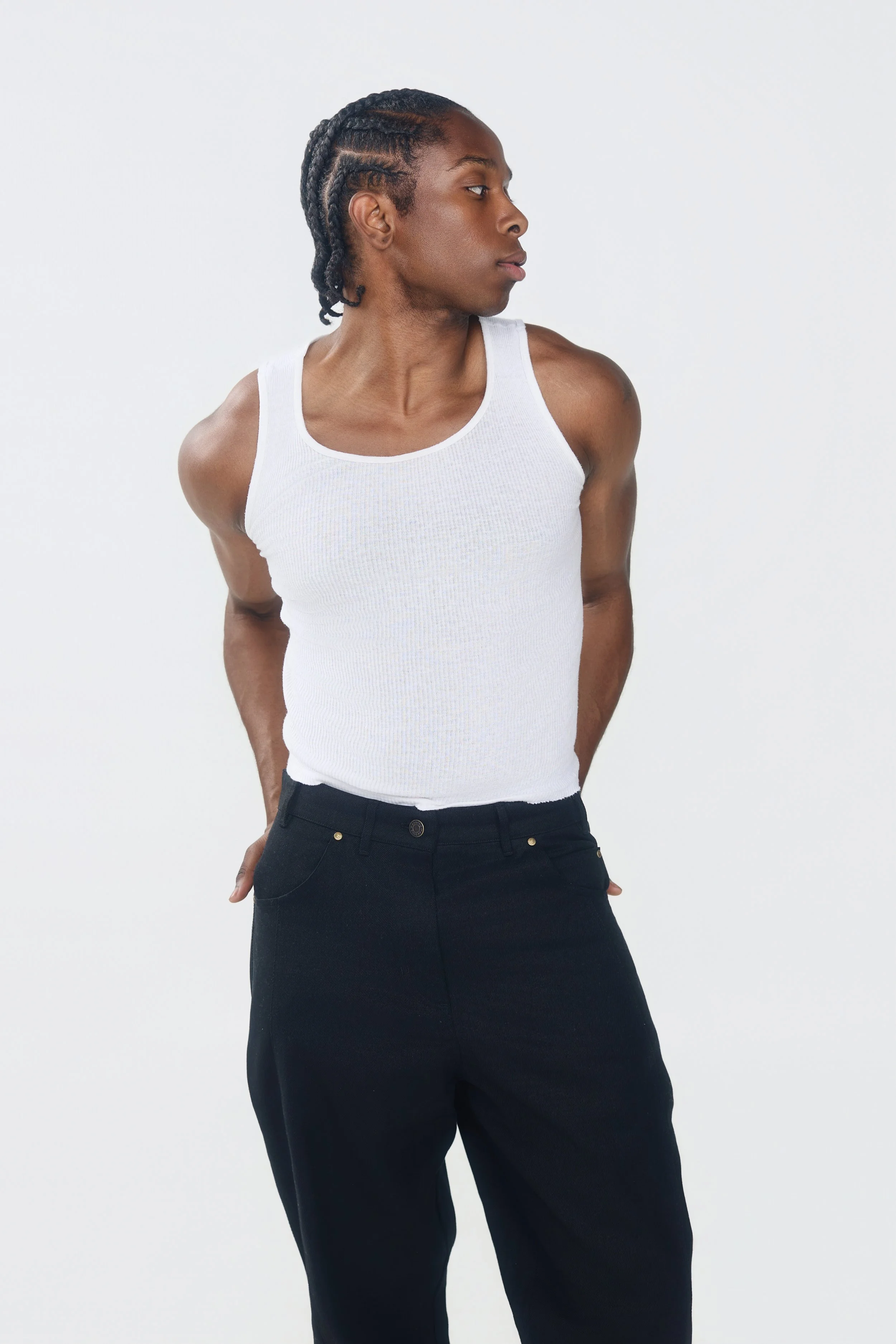 Jeune homme aux cheveux tressés portant un débardeur blanc et un pantalon noir, regardant sur le côté, à l'intérieur contre un fond blanc.