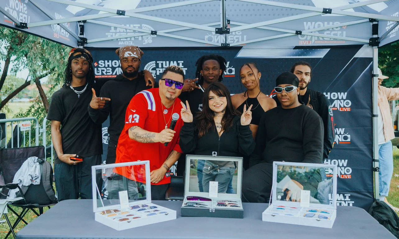 Groupe de personnes posant sous une tente lors d'un événement, avec des stands de lunettes de soleil et de montres sur une table en avant-plan.