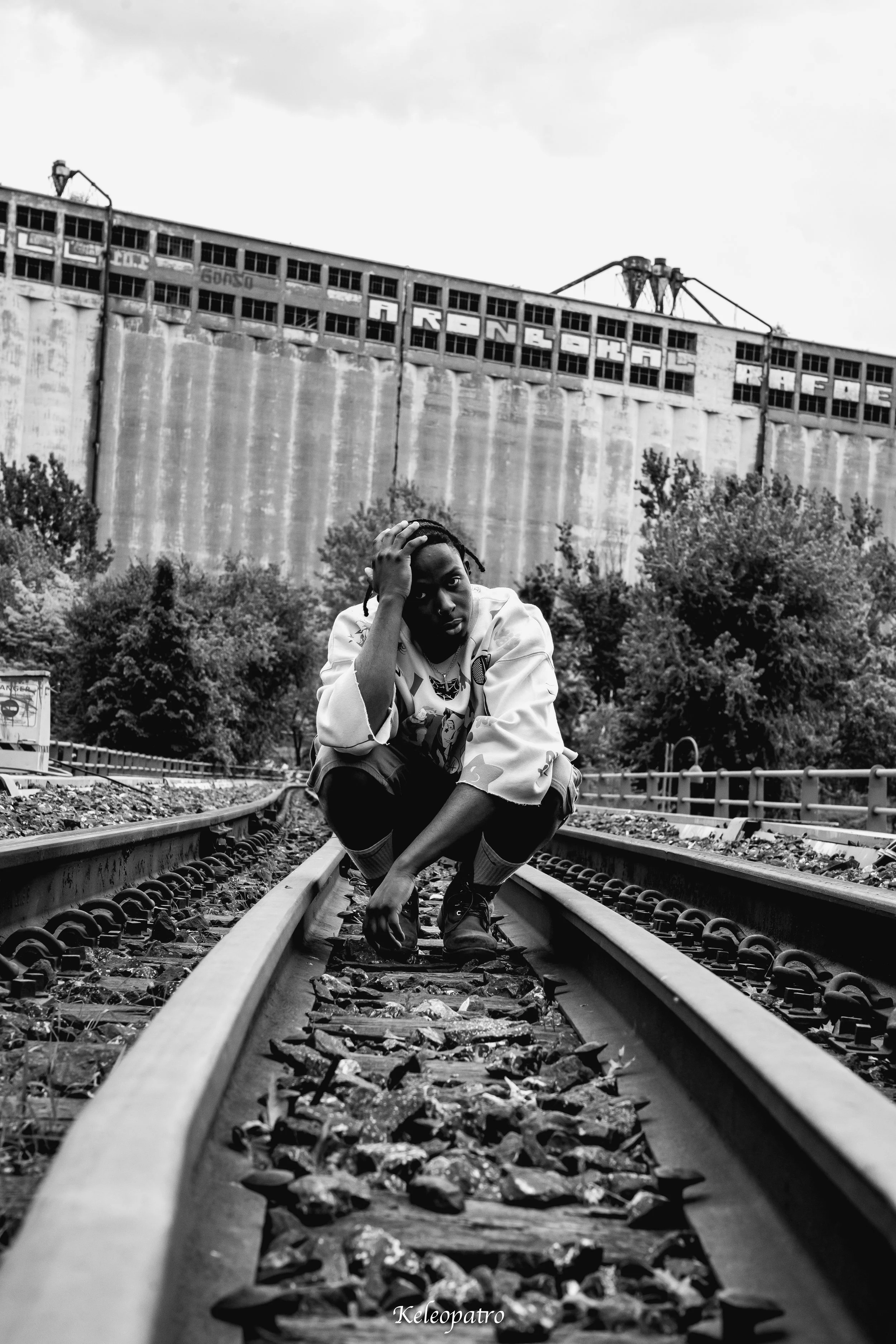Un homme assis sur des rails de chemin de fer, les genoux pliés et la tête reposant sur sa main, dans un environnement urbain avec un grand bâtiment en arrière-plan.