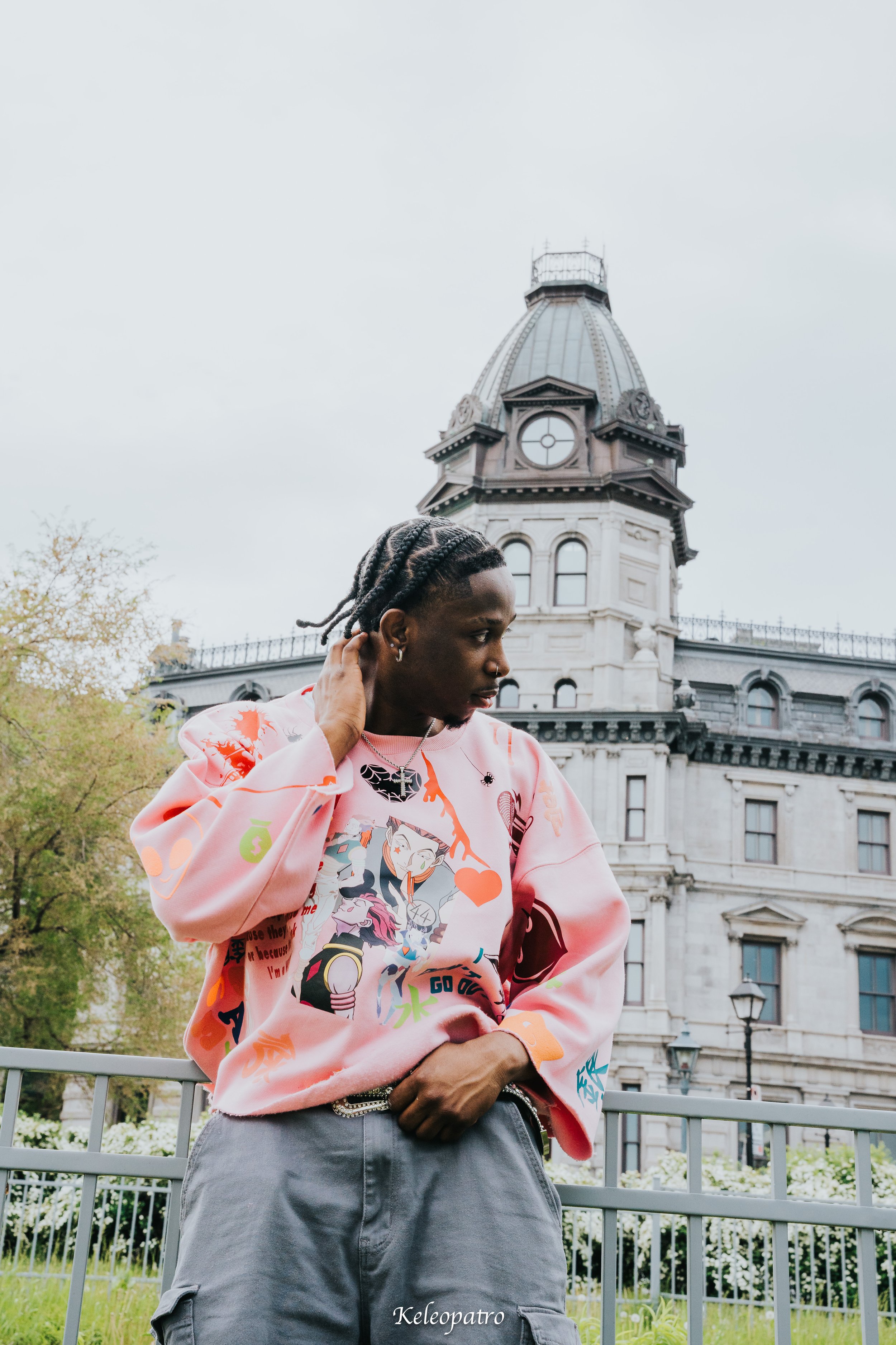 Un jeune homme avec dreadlocks portant un sweat-shirt rose avec des dessins colorés, debout dans un parc devant un bâtiment historique avec une coupole.