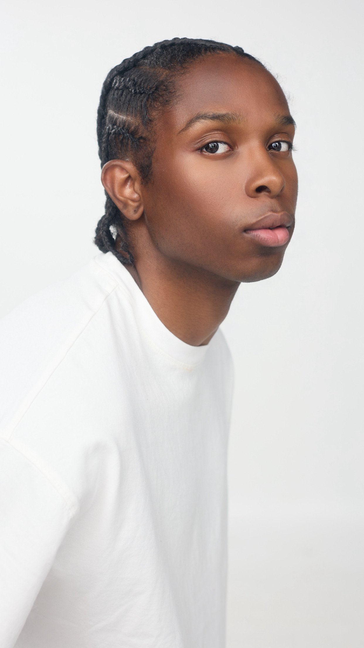 Portrait d'un jeune homme avec des cheveux tressés, portant un t-shirt blanc, contre un fond blanc.