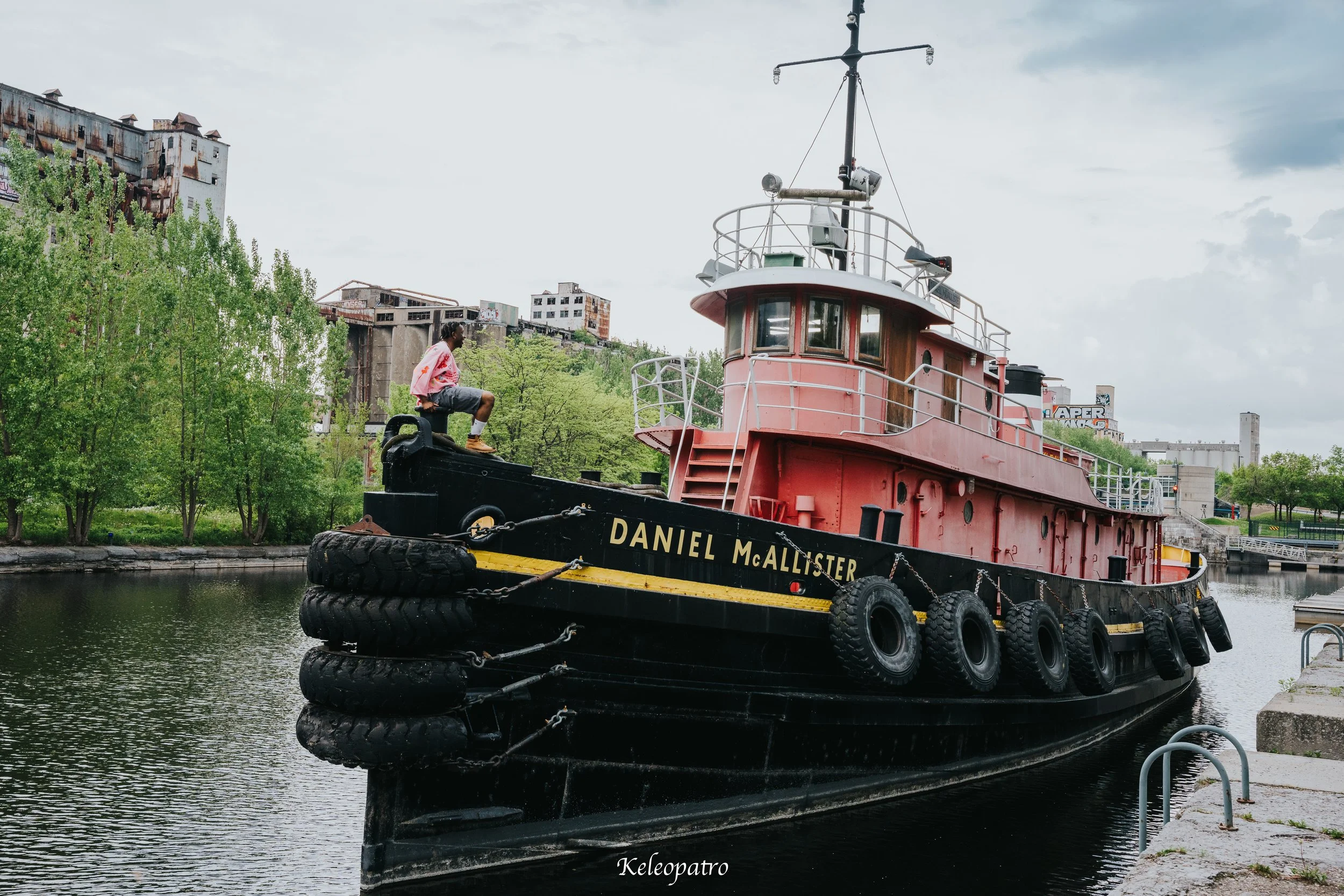 Un bateau nommé Daniel McAllister amarré dans un canal urbain avec un homme assis sur la proue, entouré d'arbres verts et de bâtiments en arrière-plan, sous un ciel nuageux.