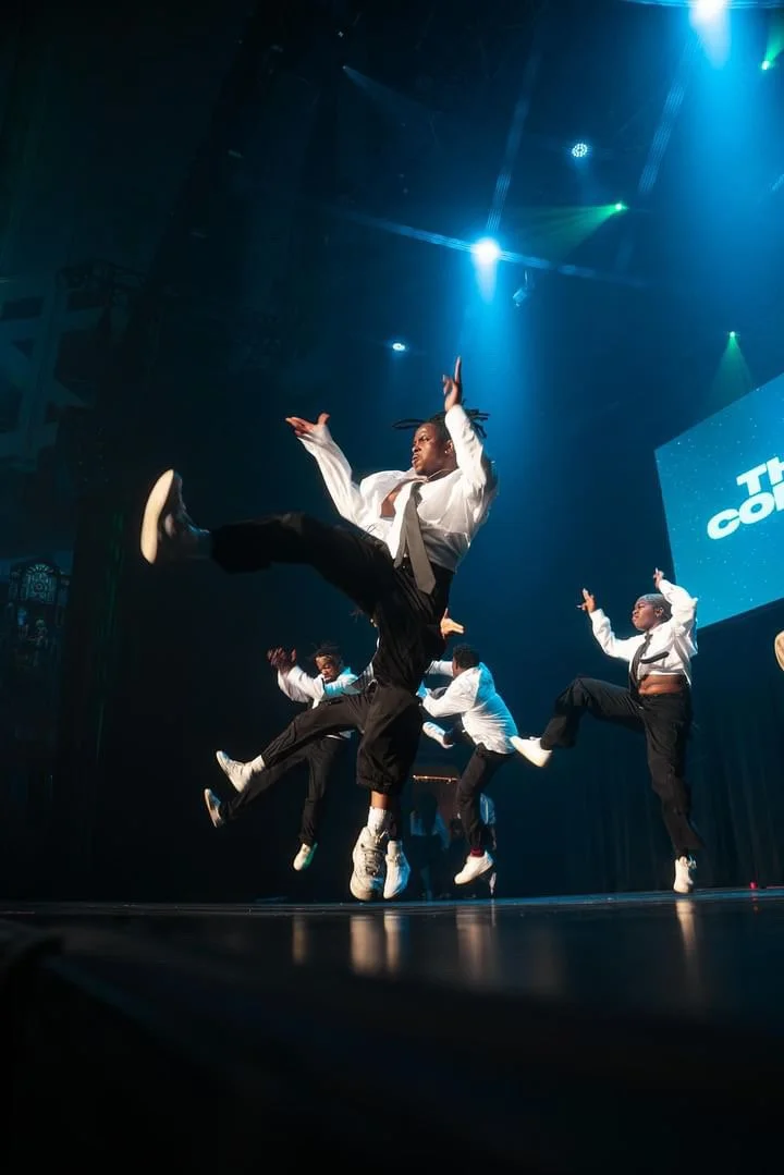 Groupe de danseurs effectuant une chorégraphie sur scène sous des lumières colorées.