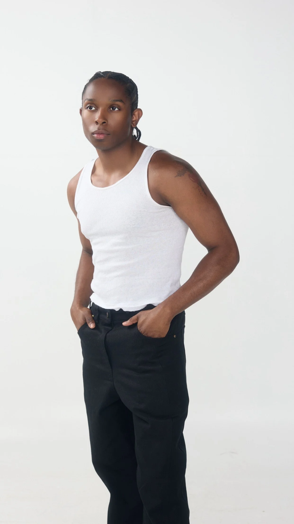 Jeune personne portant un débardeur blanc et un pantalon noir, positionnée contre un fond blanc, regardant à gauche, mains dans les poches.