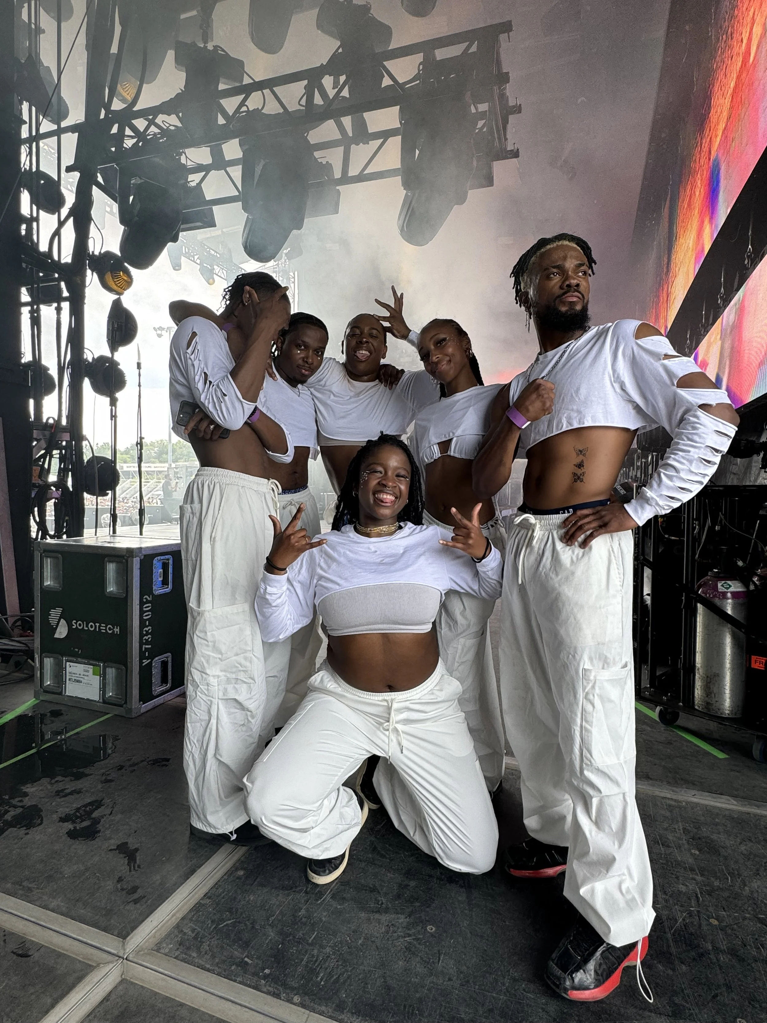 Groupe de six personnes en tenues décontractées blanches, posant et souriant sur scène avec éclairage et équipements de concert en arrière-plan.