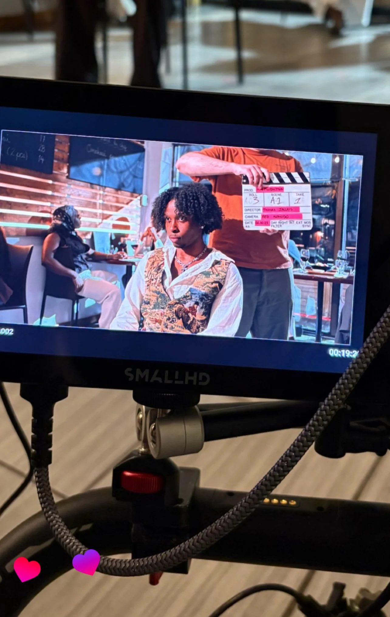 A scene from a film or video shoot showing a young woman with curly hair seated indoors, with a crew member holding a clapperboard behind her. The setting appears to be a restaurant or cafe with people in the background, including a woman sitting on a couch. The scene is being filmed with professional equipment, including a monitor in the foreground.