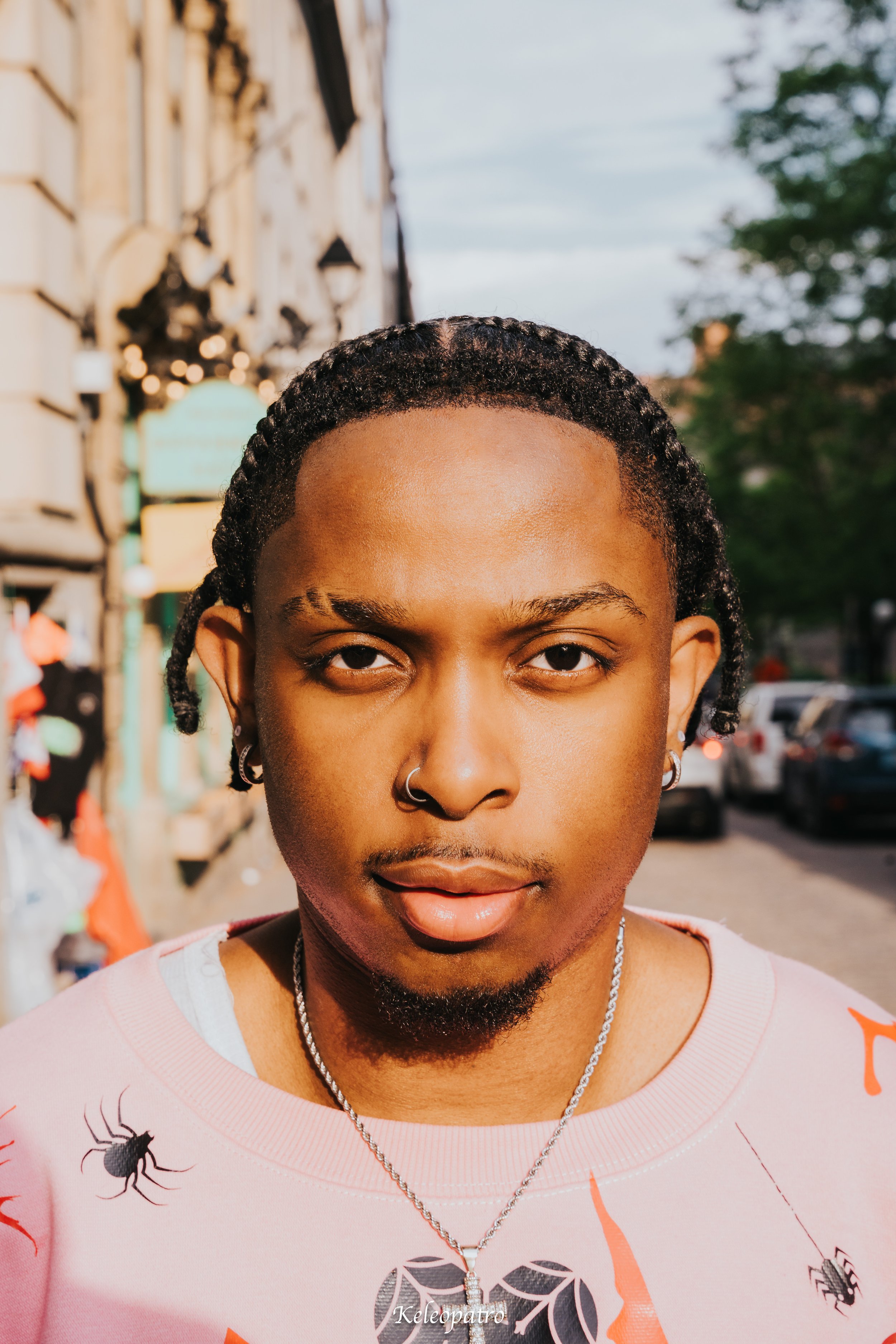 A young person with short curly hair and earrings, standing outdoors on a city street with cars and trees in the background.