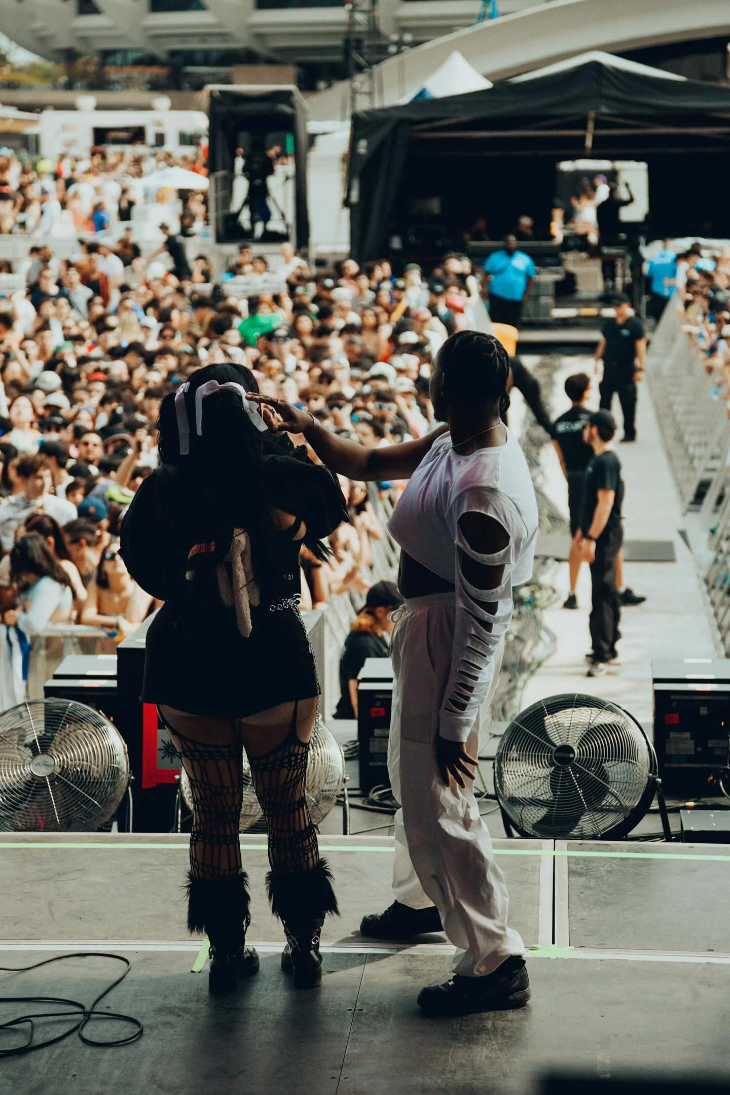 Deux personnes côte à côte sur une scène lors d'un concert ou d'un festival. L'une porte une tenue noire avec des vêtements déchirés et des chaussettes en laine haute, l'autre porte une tenue blanche avec des manches déchirées. En arrière-plan, il y a une grande foule assise et debout avec une scène principale, des enceintes, un écran et des personnes en train de surveiller la scène.