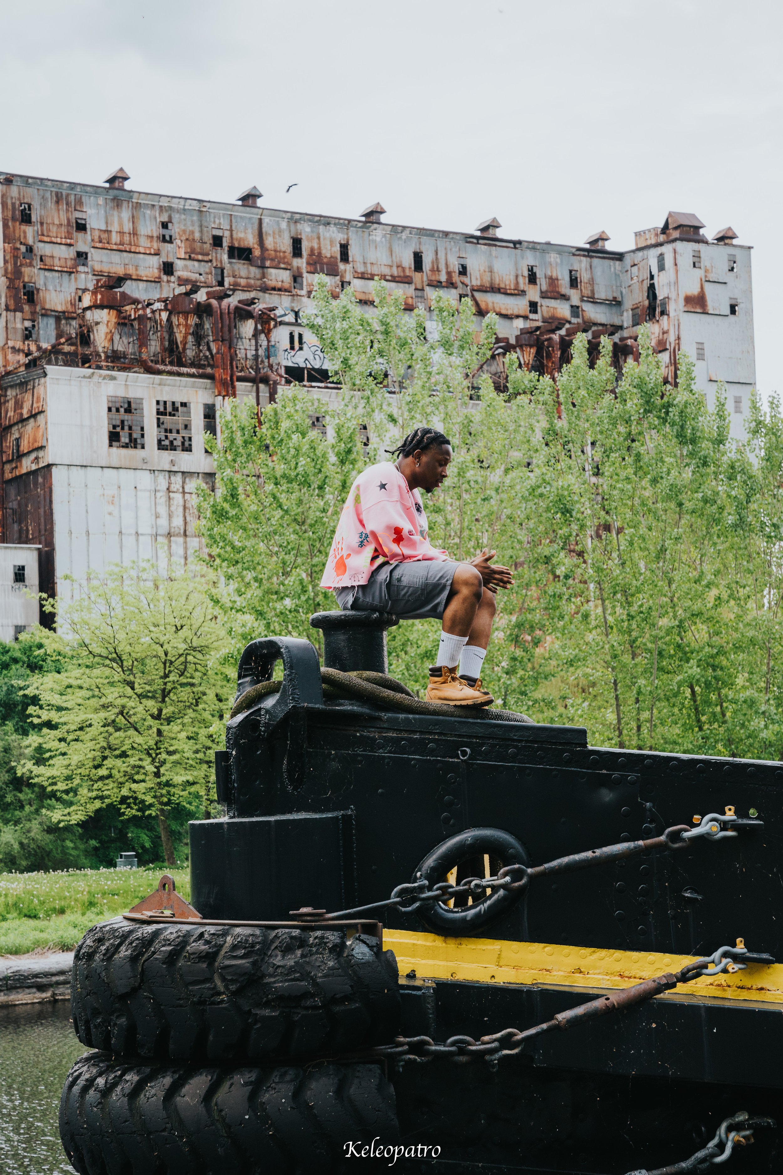 Un homme assis sur une plateforme noire, muni de vêtements de style urbain, regarde vers le bas, avec un bâtiment en ruine derrière lui et un arbre vert à l'avant-plan.