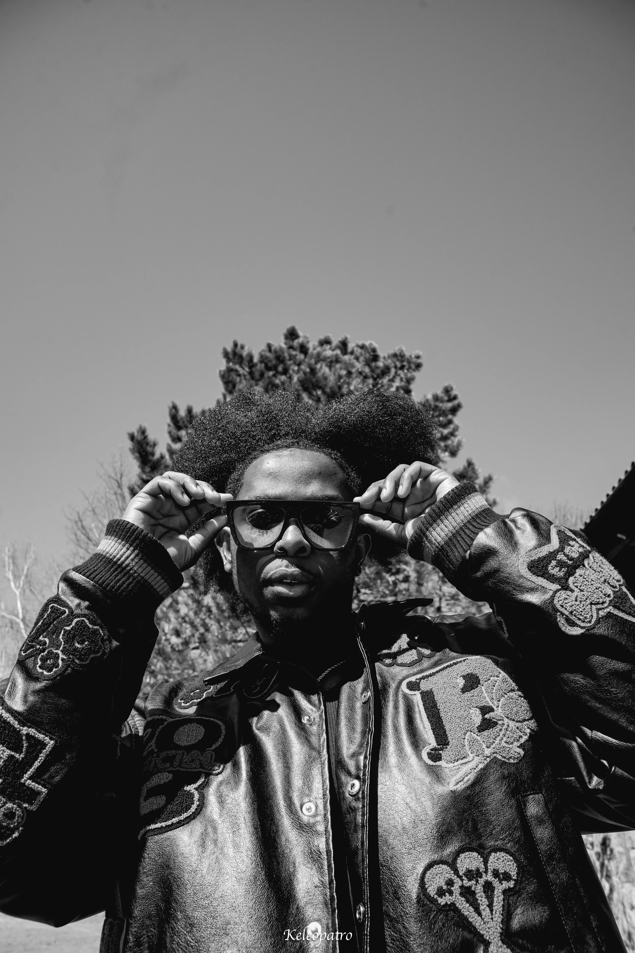 Un homme portant des lunettes de soleil et une veste en cuir avec des patchs, debout à l'extérieur, avec des arbres en arrière-plan, dans une image en noir et blanc.