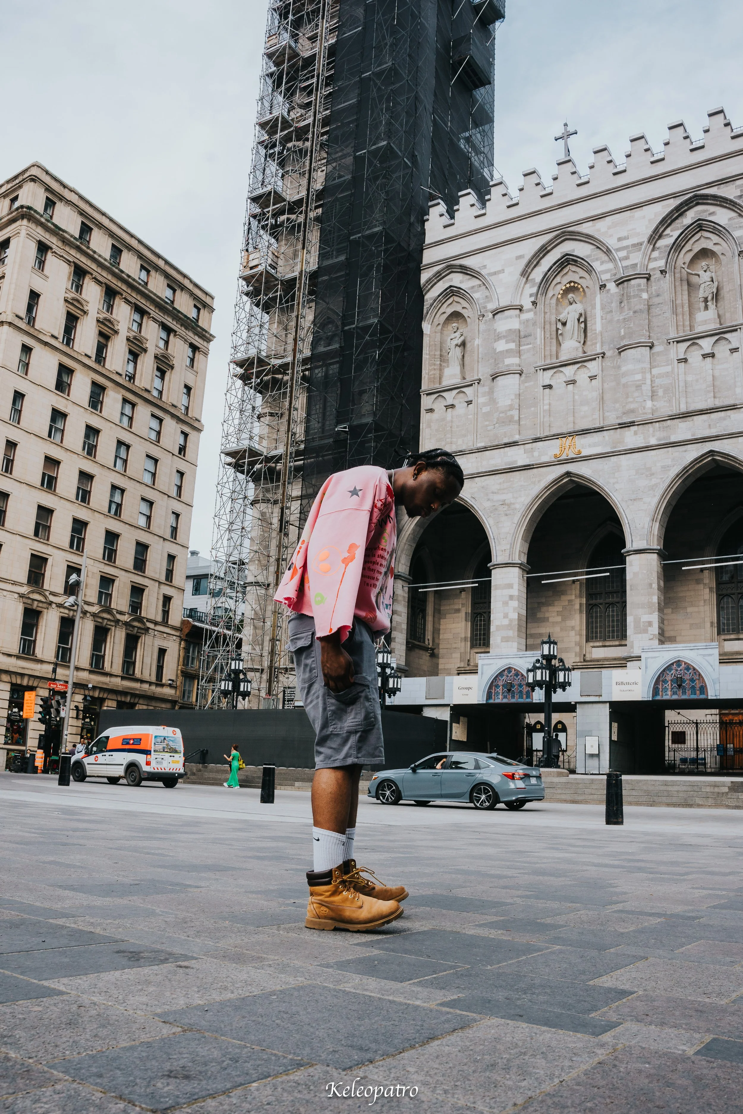 A young man standing on a city street crossing with his head bowed. He is dressed in a pink hoodie with colorful designs, gray shorts, white socks, and tan boots. In the background, there is a large historical building with Gothic architecture and sc