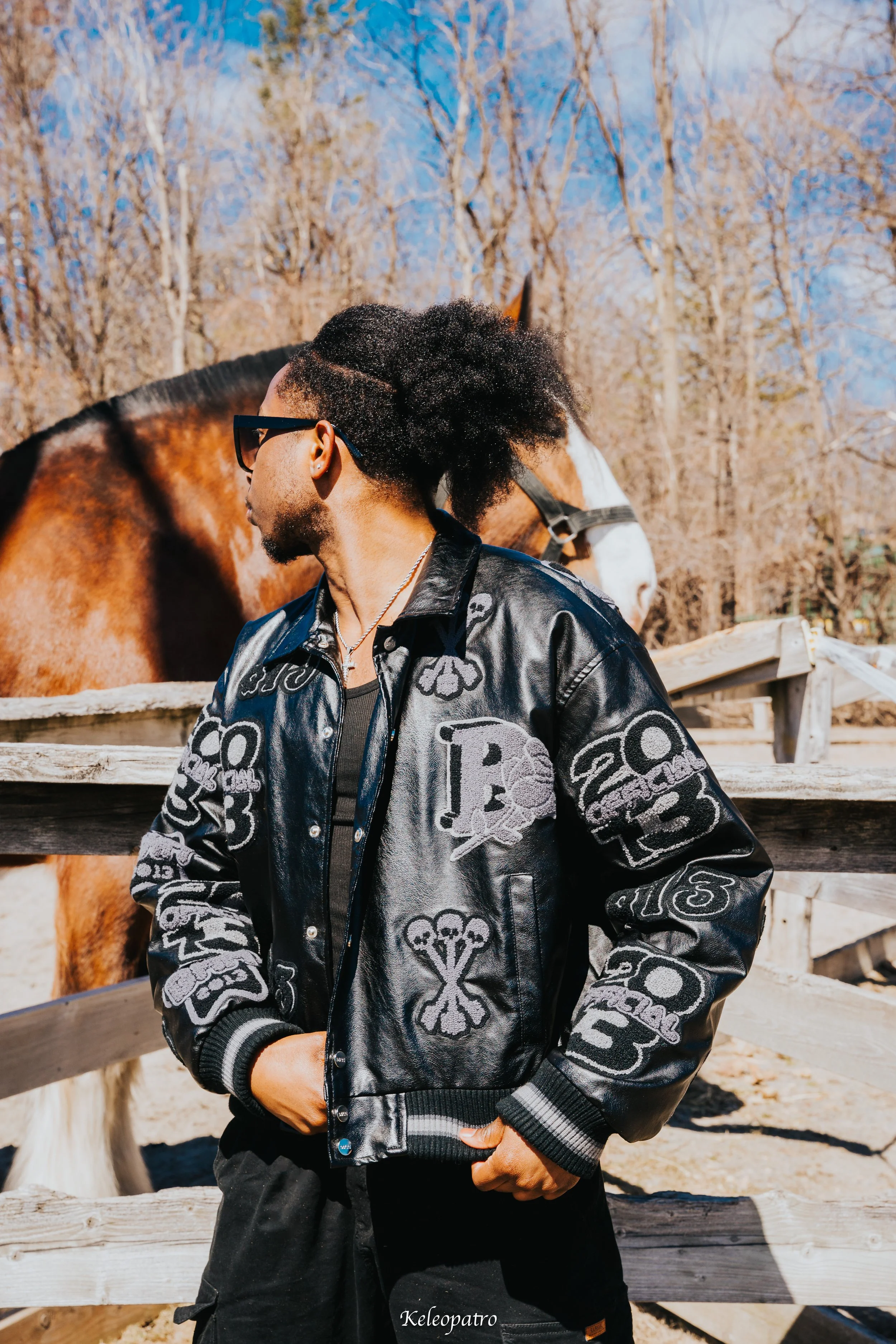 Jeune homme portant une veste en cuir noire avec des motifs, portant des lunettes de soleil, debout à côté d'une clôture en bois, avec un cheval en arrière-plan dans un décor extérieur avec des arbres et un ciel bleu.