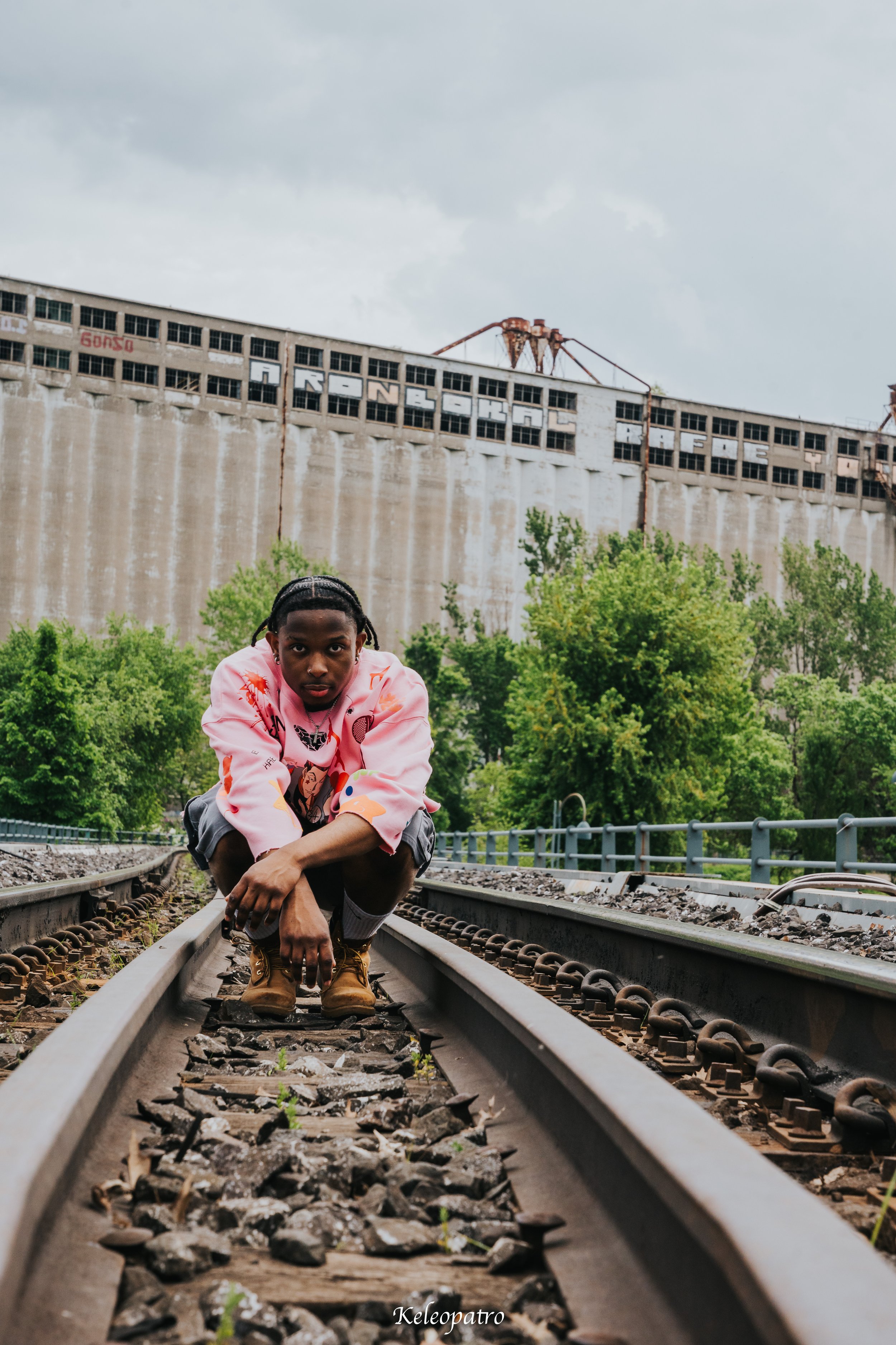 Un jeune homme en sweatshirt rose et chaussures marron, squattant sur des rails de train, avec un fond de bâtiment industriel et des arbres verts.