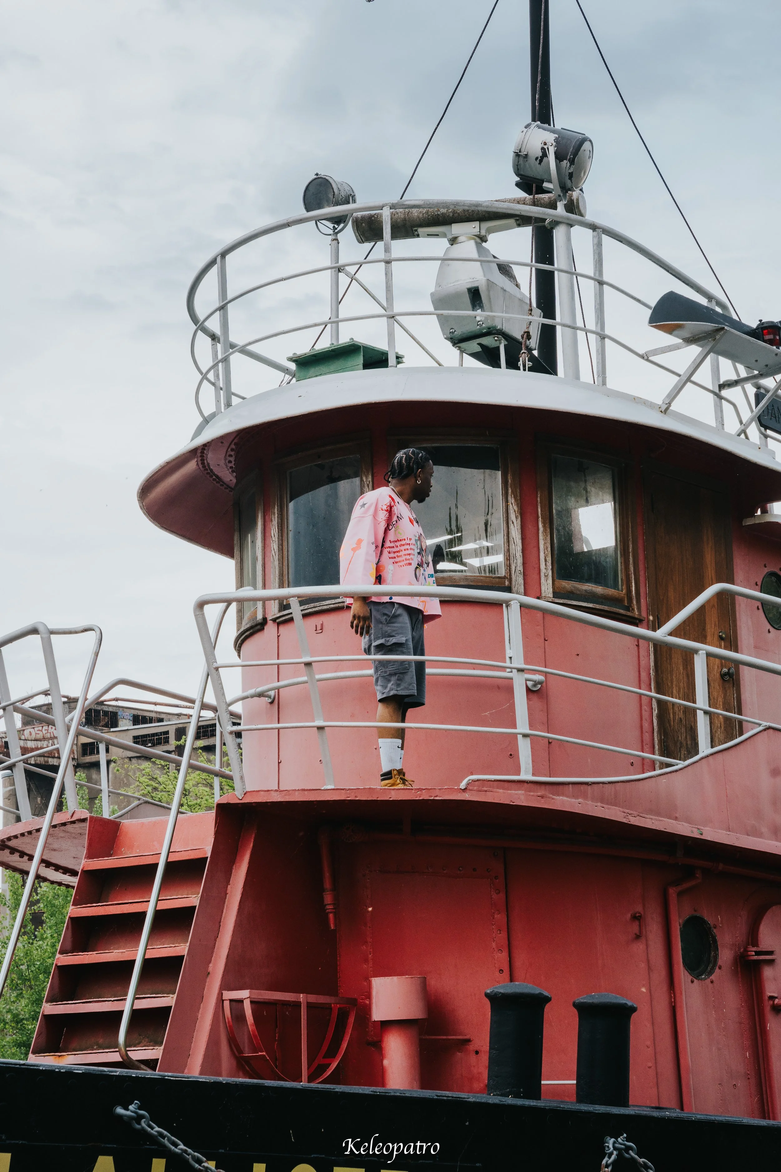 Une personne debout sur la plateforme d'un vieux bateau rouge, regardant vers la gauche, avec un ciel nuageux en arrière-plan.