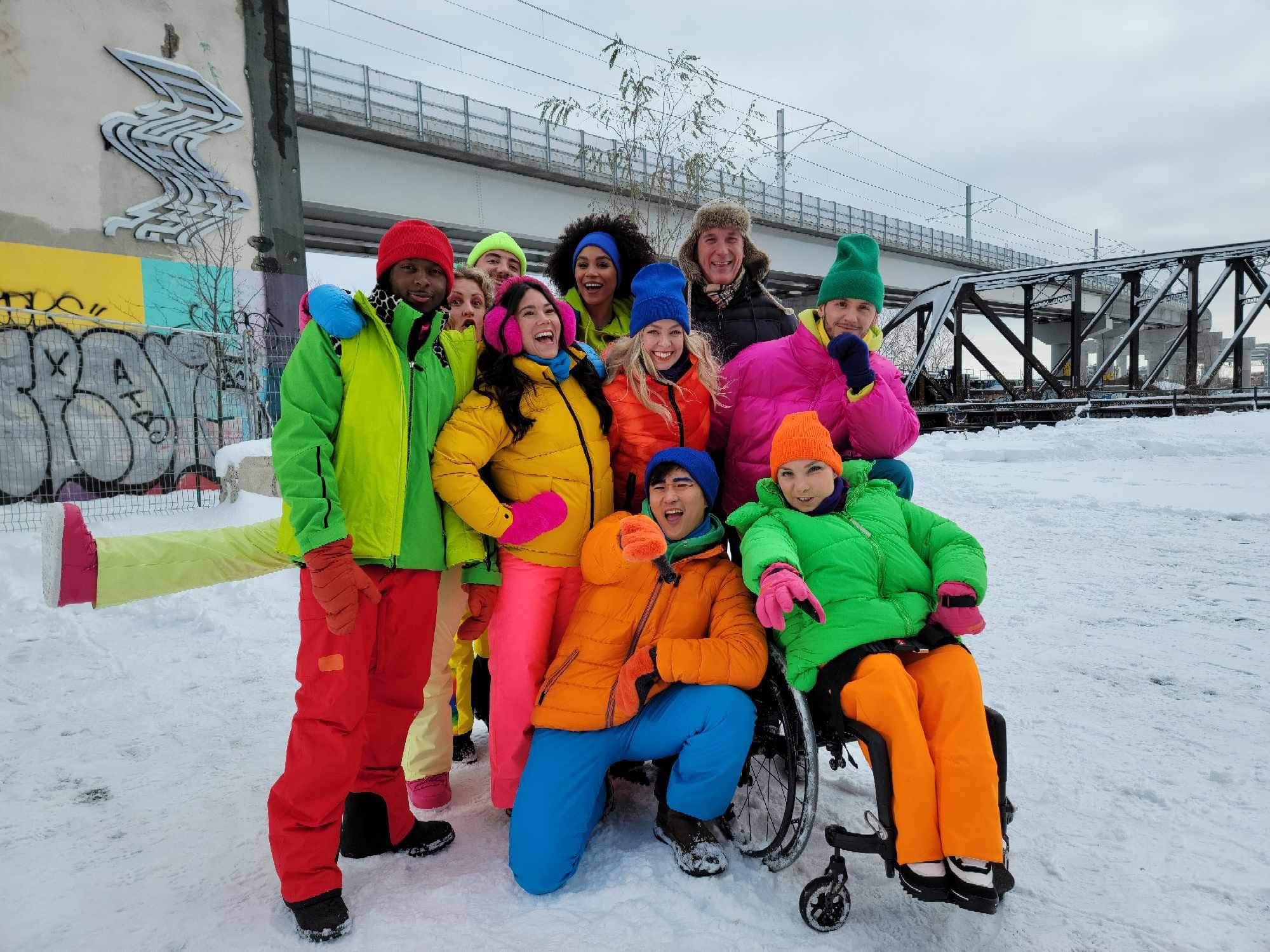 Groupe diversifié de personnes posant dans la neige, portant des vêtements d'hiver colorés, avec un pont et un mur graffité en arrière-plan.