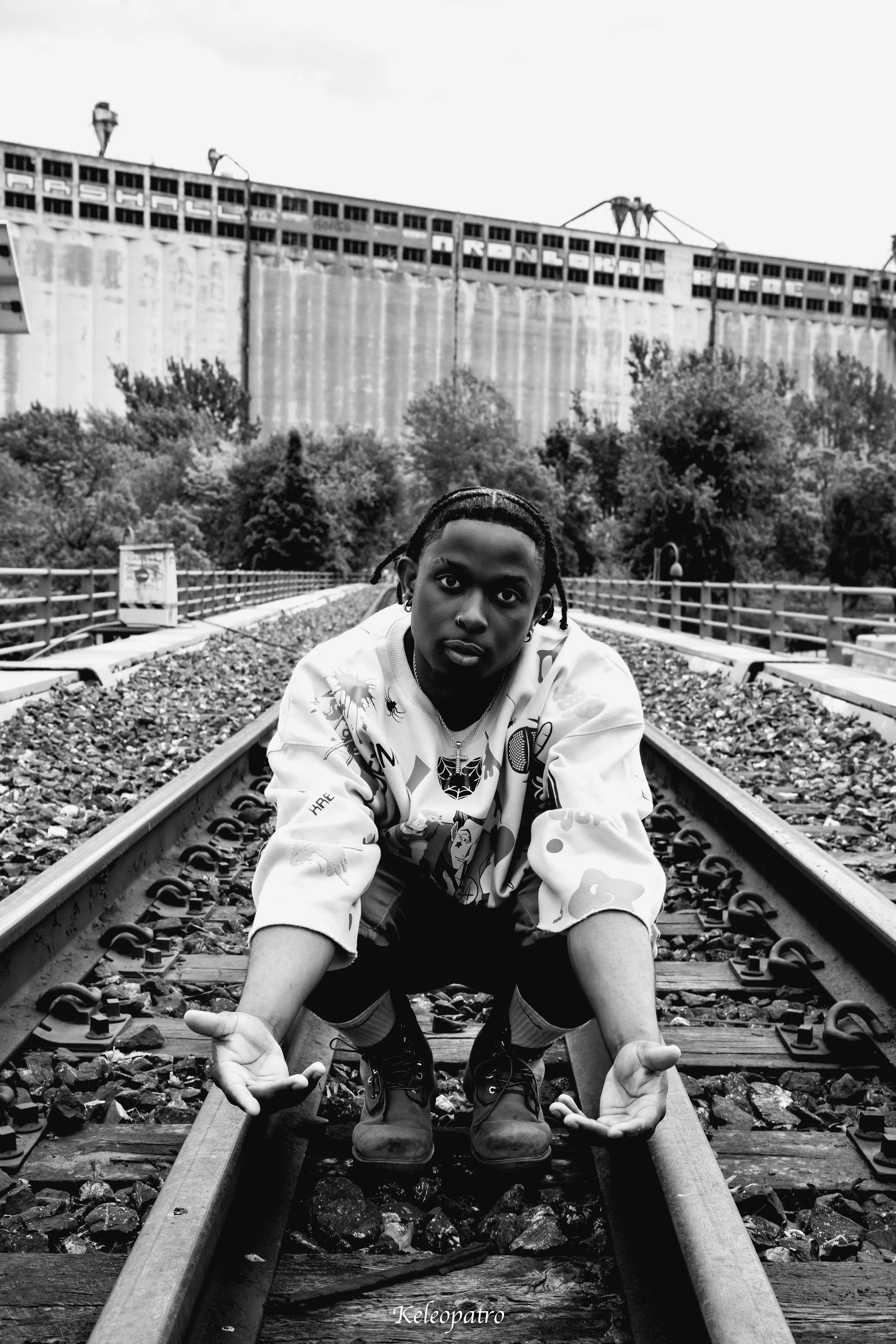 Person crouching on train tracks in an outdoor setting, with trees and an industrial building in the background, in black and white.