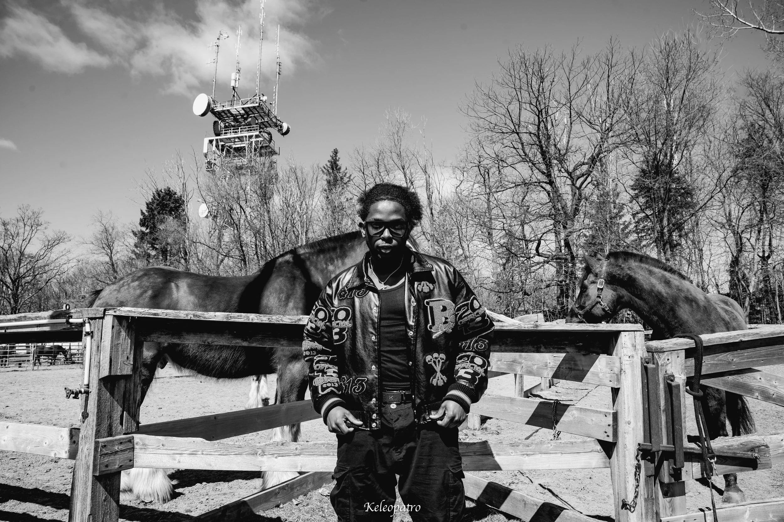 Un jeune homme portant des lunettes noires, un blouson en cuir avec des motifs, se tient devant une clôture en bois dans un enclos avec deux chevaux bruns, avec des arbres dénudés en arrière-plan et une tour de communication en position élevée dans l