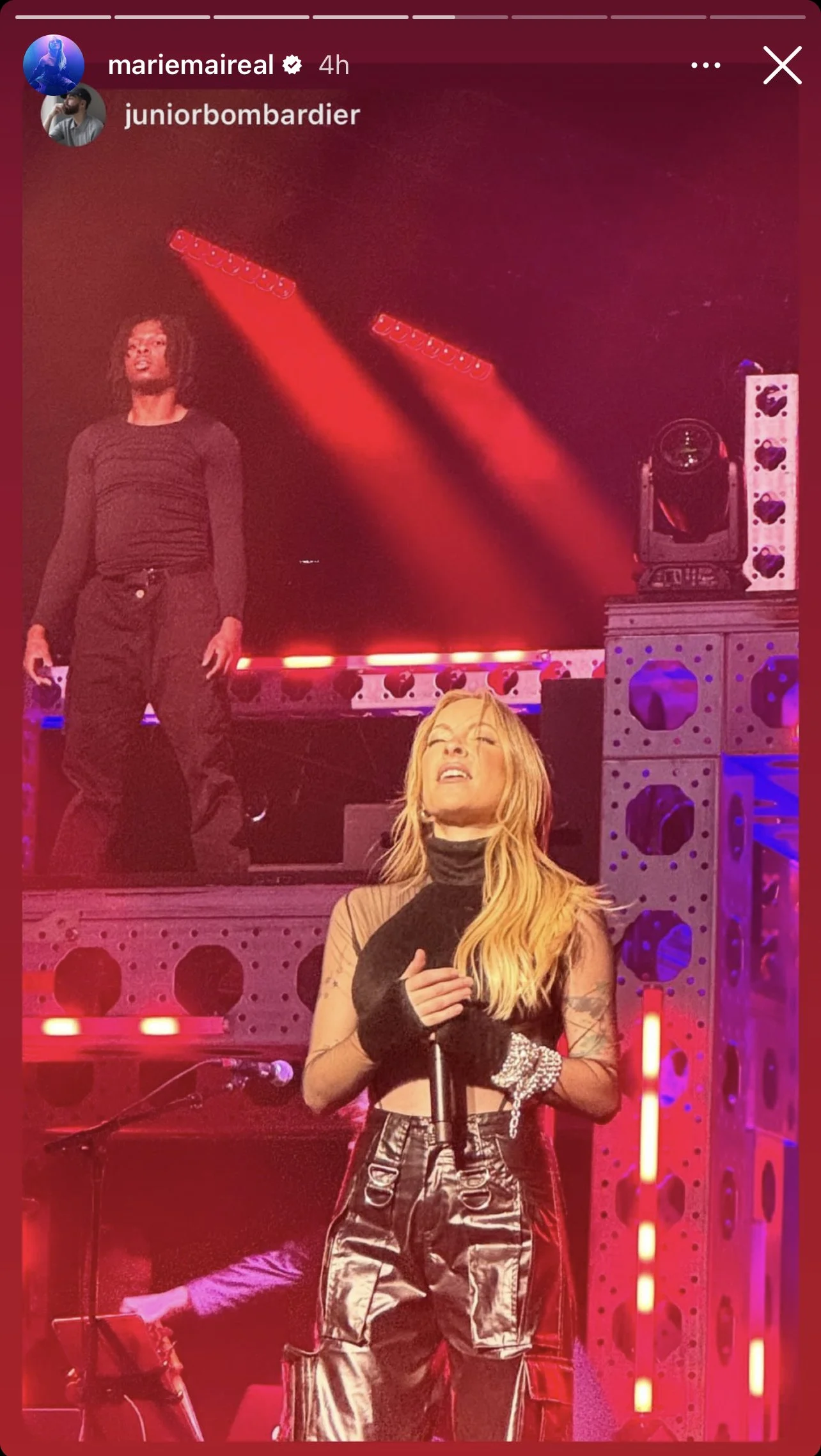 Une femme chante lors d'un concert sur scène, avec des lumières rouges et un homme derrière elle, debout. La scène est équipée d'éclairages et d'enceintes.