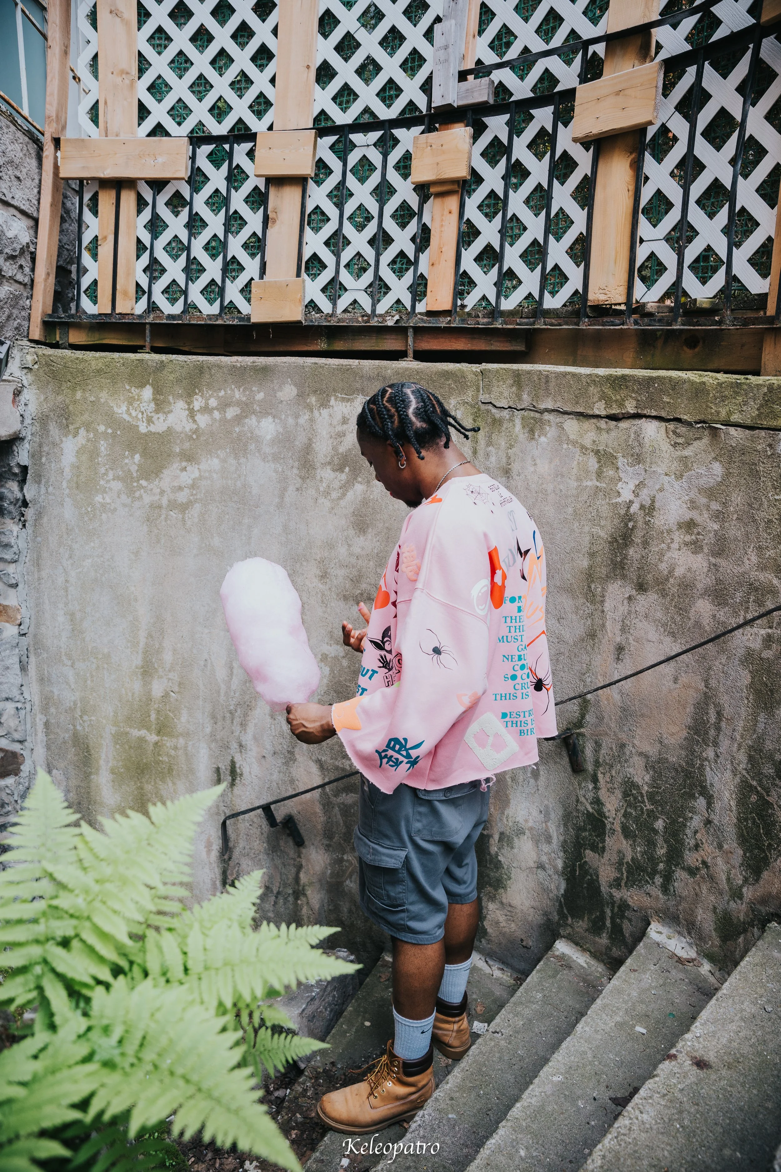Un jeune homme debout, regardant une petite boule de coton ou une barbe à papa rose, dans une cour extérieure avec un mur en béton et une structure en bois et treillis blanc en arrière-plan, portant une veste rose avec des motifs colorés.
