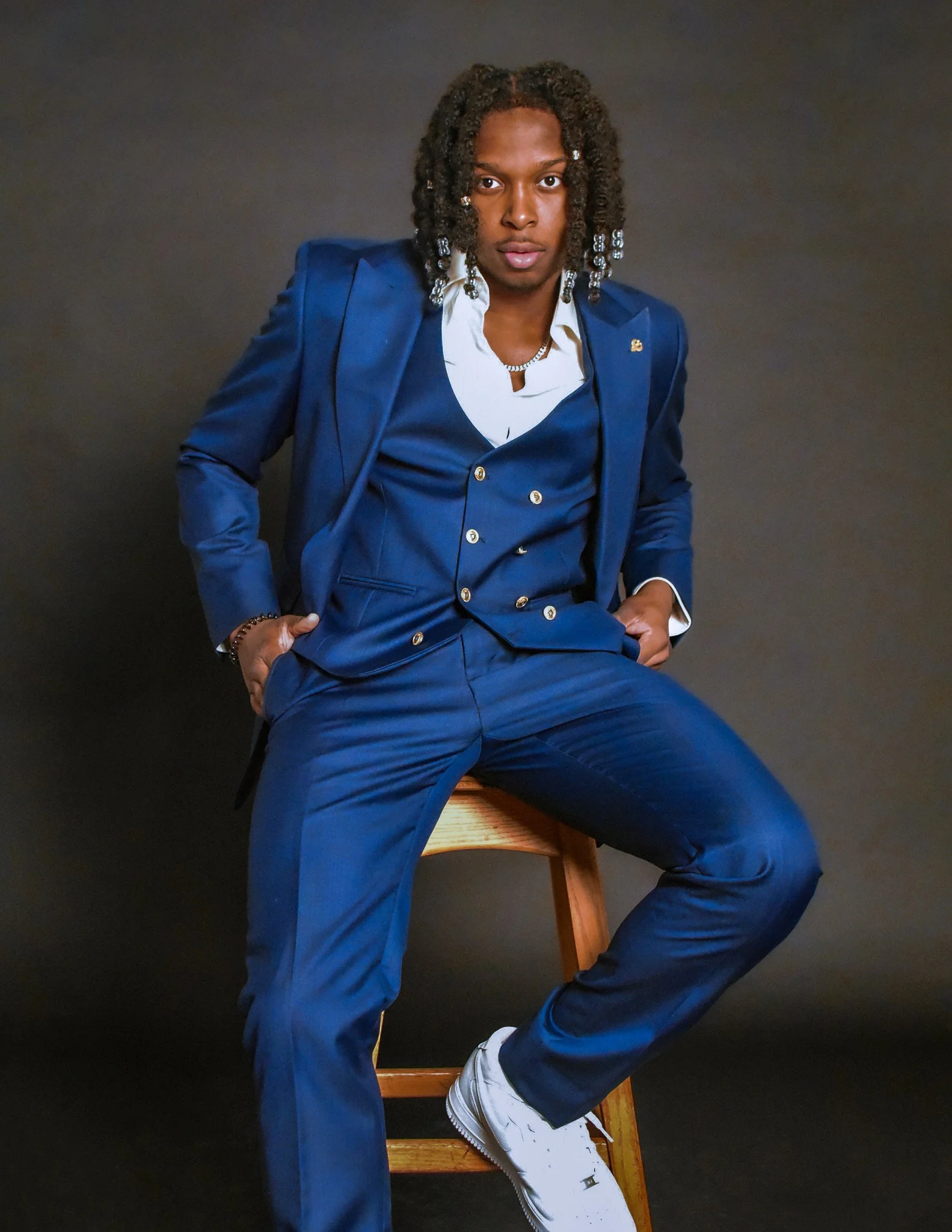 Homme noir portant un costume bleu avec une chemise blanche, assis sur une chaise en bois contre un fond sombre.