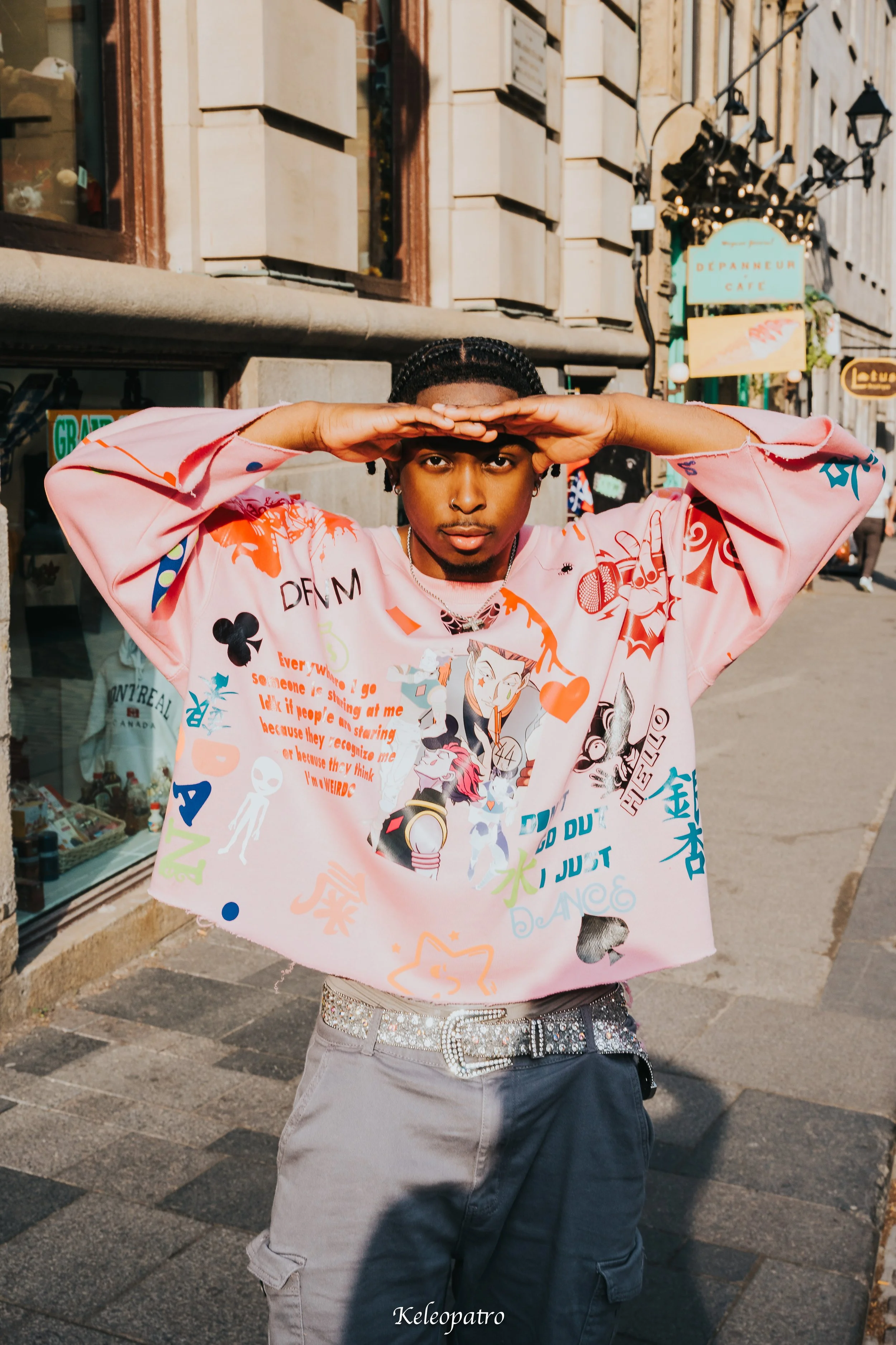 A young man standing on a city sidewalk, wearing a colorful graphic sweatshirt and gray pants, poses with hands shielding his eyes from the sun. There are storefronts and street signs in the background.
