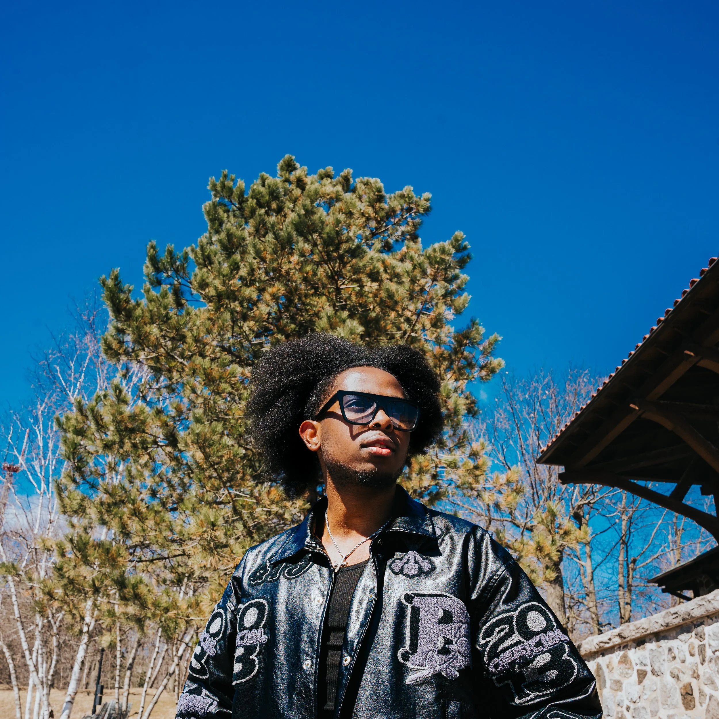 A young man with large sunglasses and an afro hairstyle standing outdoors against a backdrop of a tree with green foliage and a clear blue sky.