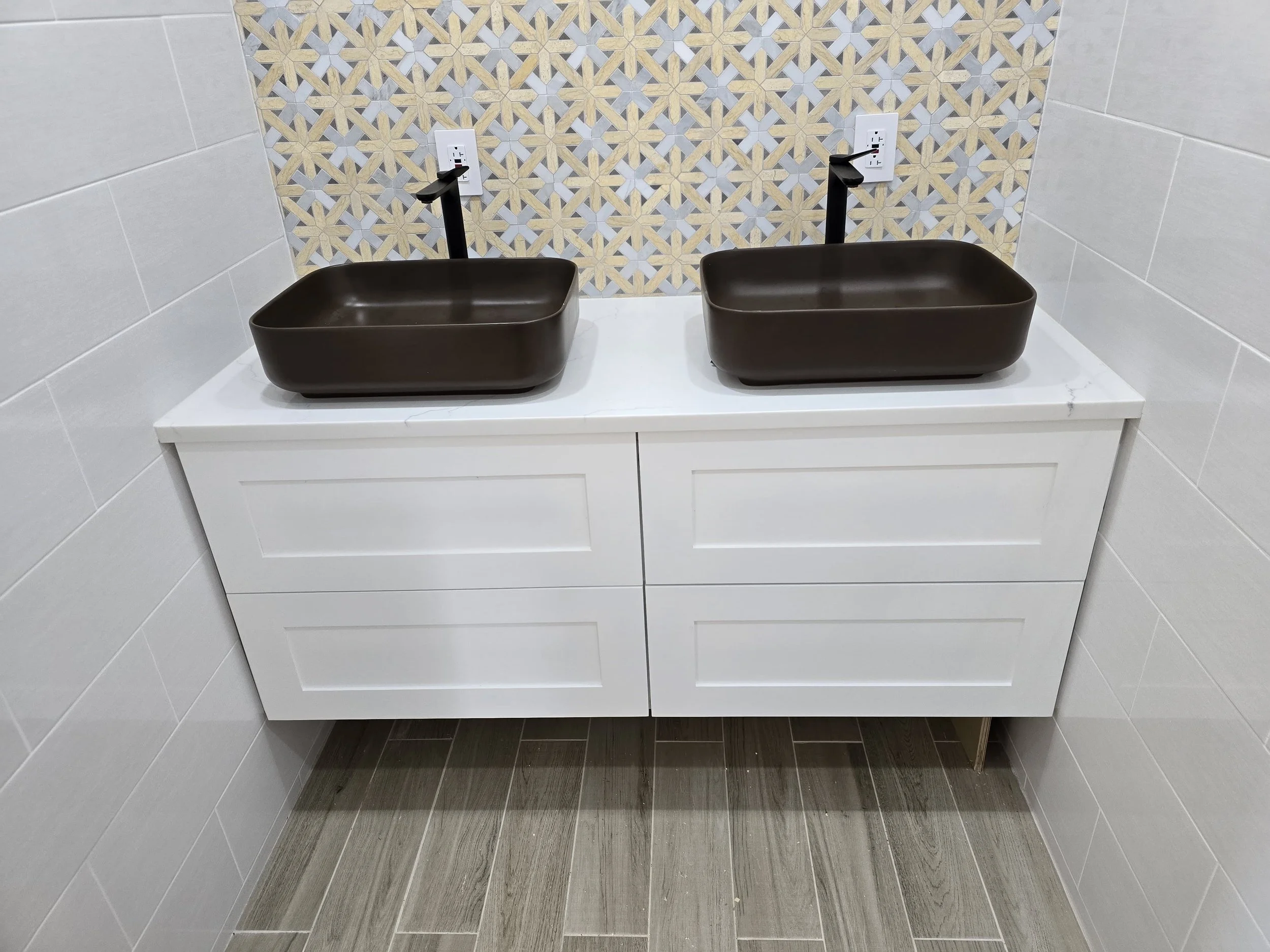 A bathroom double vanity with two black rectangular vessel sinks and black faucets mounted on a white cabinet beneath a tiled wall with beige, cream, and gray pattern.