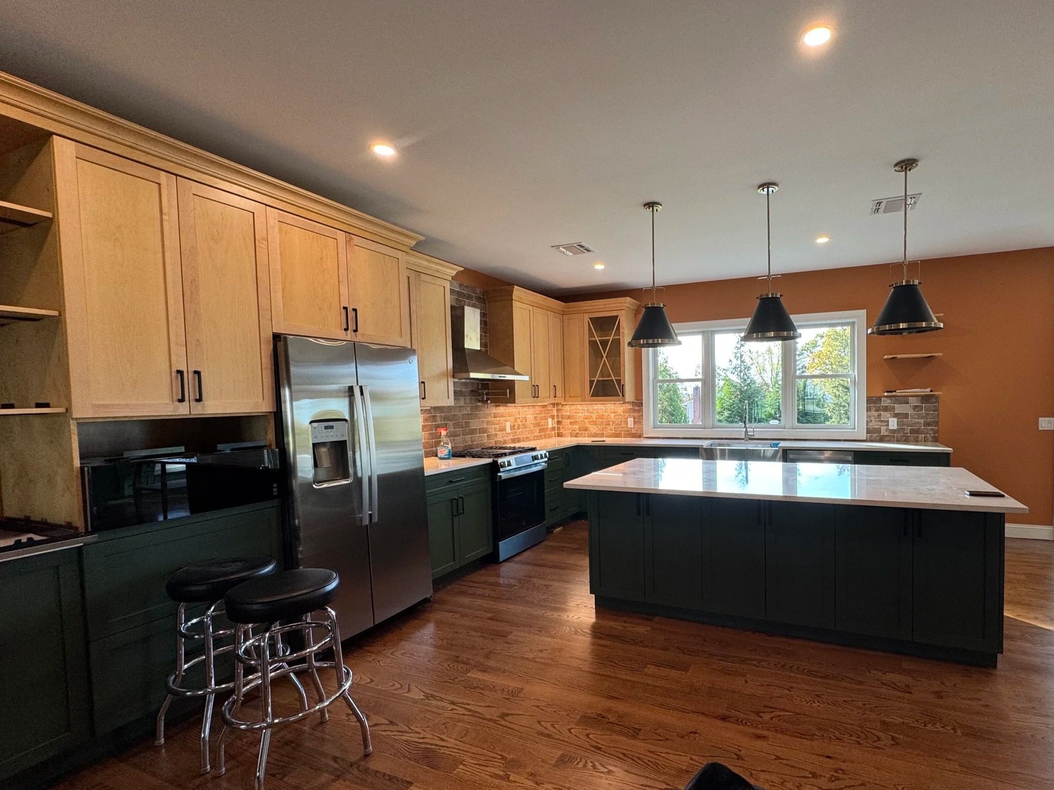 Modern kitchen with dark green lower cabinets, light wood upper cabinets, brick backsplash, large kitchen island with a marble countertop, stainless steel refrigerator, and black pendant lights above the island. There are windows at the far end of th