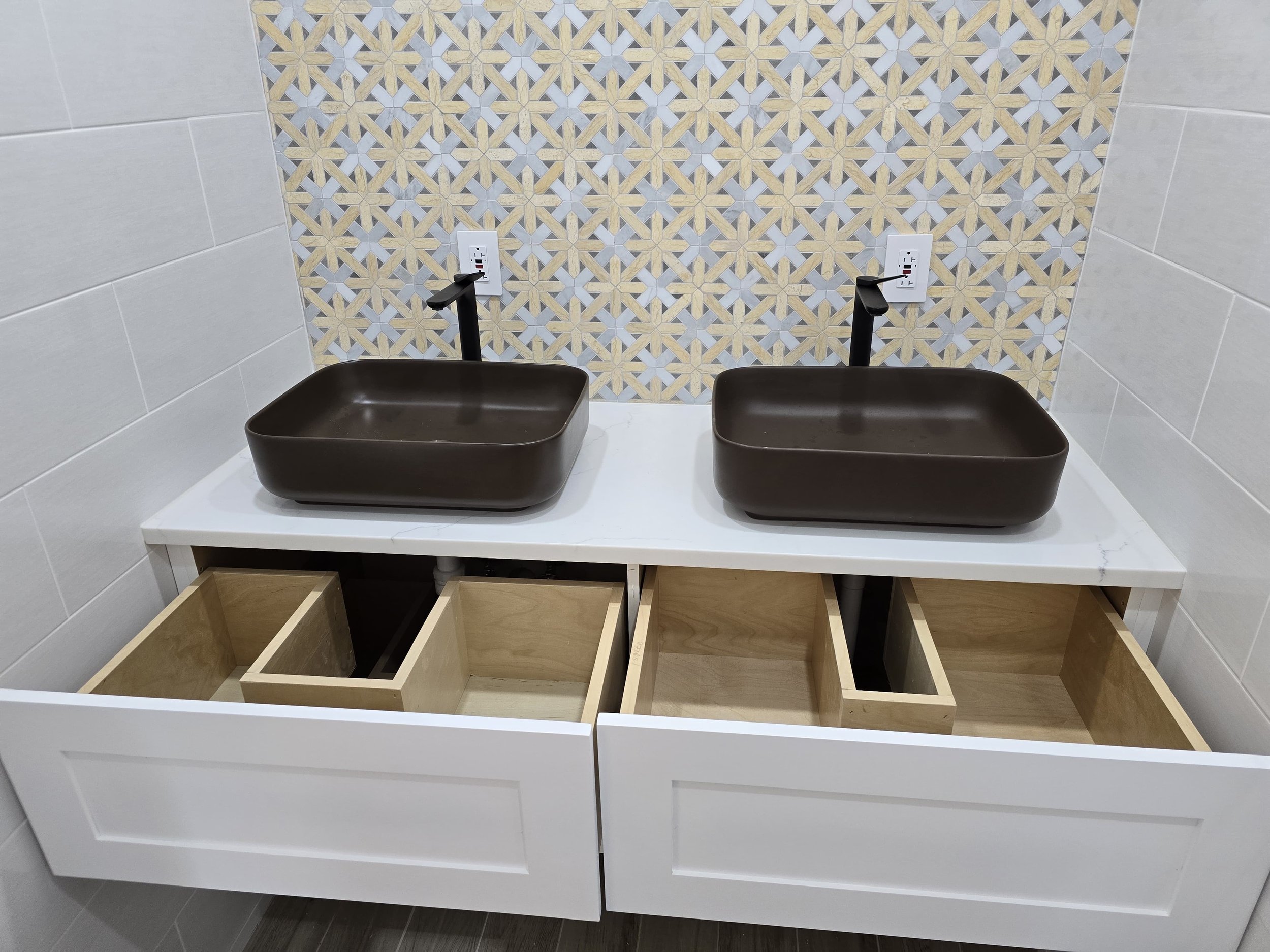 Double bathroom vanity with two brown vessel sinks and black faucets, open white cabinet with empty wooden drawers, and a decorative tile backsplash