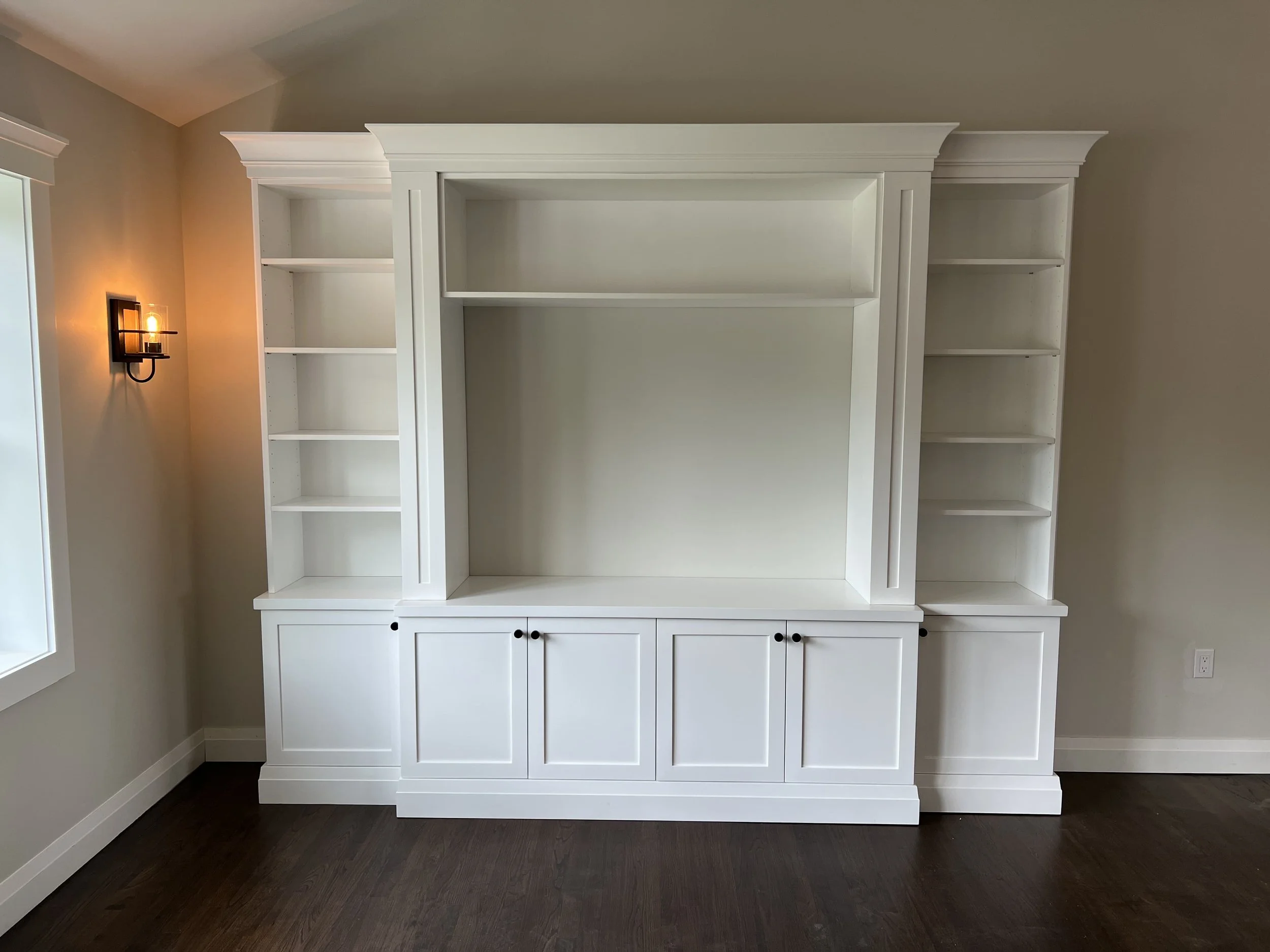 White built-in shelving unit with cabinets at the bottom, set against a beige wall with a wall-mounted light fixture on the left side, and a window with white trim on the far left.