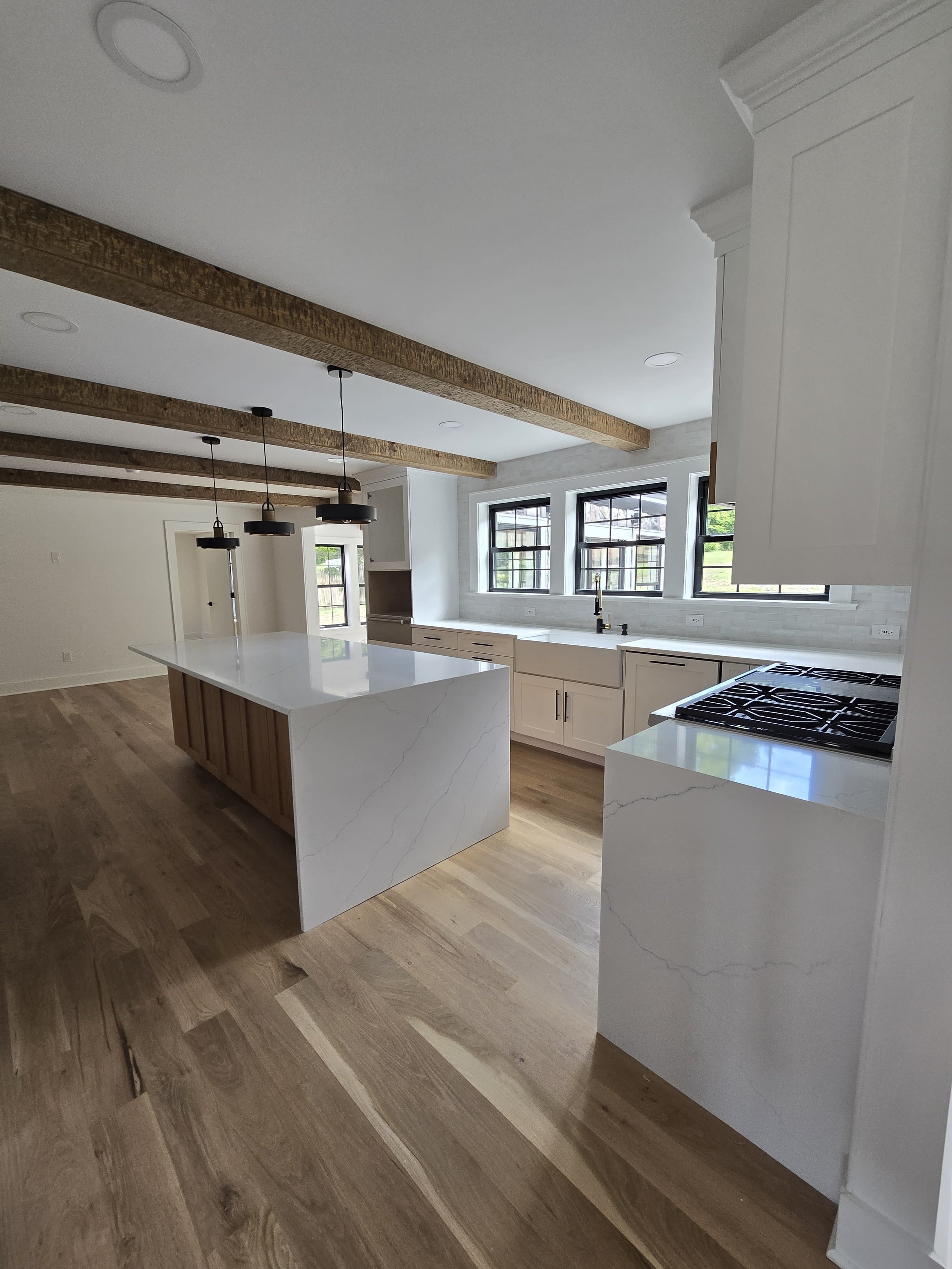 Modern kitchen with wooden beams on the ceiling, white cabinets, a large island with a white marble countertop, three black pendant lights, and three large windows with black frames overlooking a green outdoor area.
