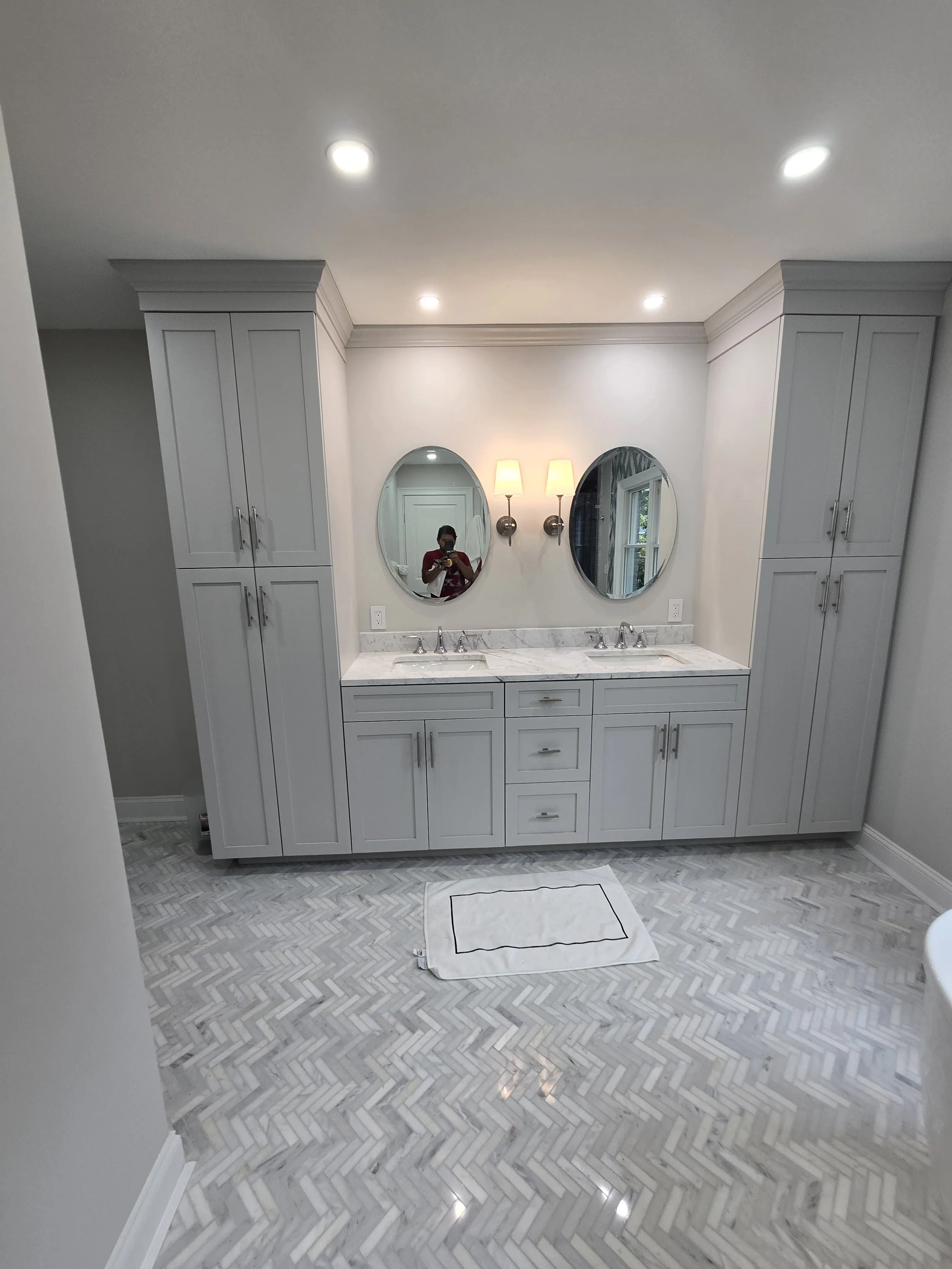 Modern bathroom with double vanity, marble countertops, two oval mirrors, wall-mounted light fixtures, white cabinetry, herringbone tile floor, and a white rug.