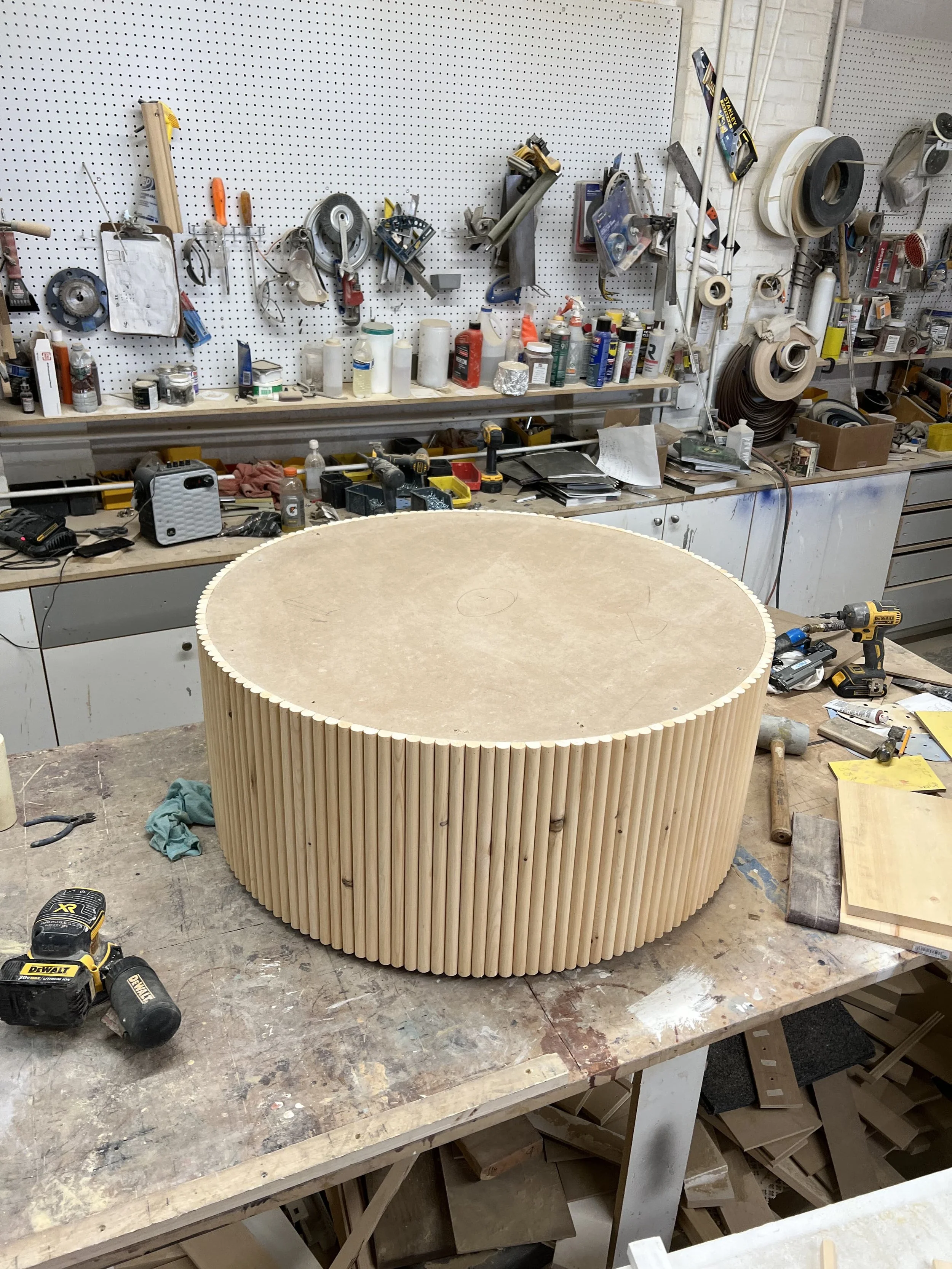 A woodworking workshop with a large circular piece of wood with vertical wooden dowels around it on a workbench, surrounded by tools, drill, and work supplies.