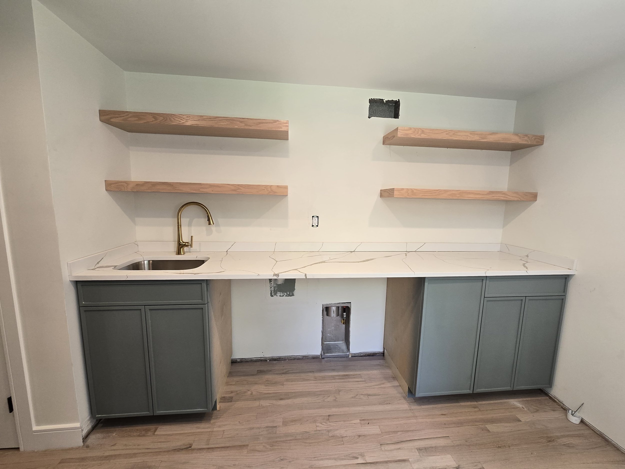 Kitchen area under renovation with a marble countertop, two green cabinets underneath, a gold faucet, and three wooden shelves on the wall. There are missing appliances and open electrical boxes.