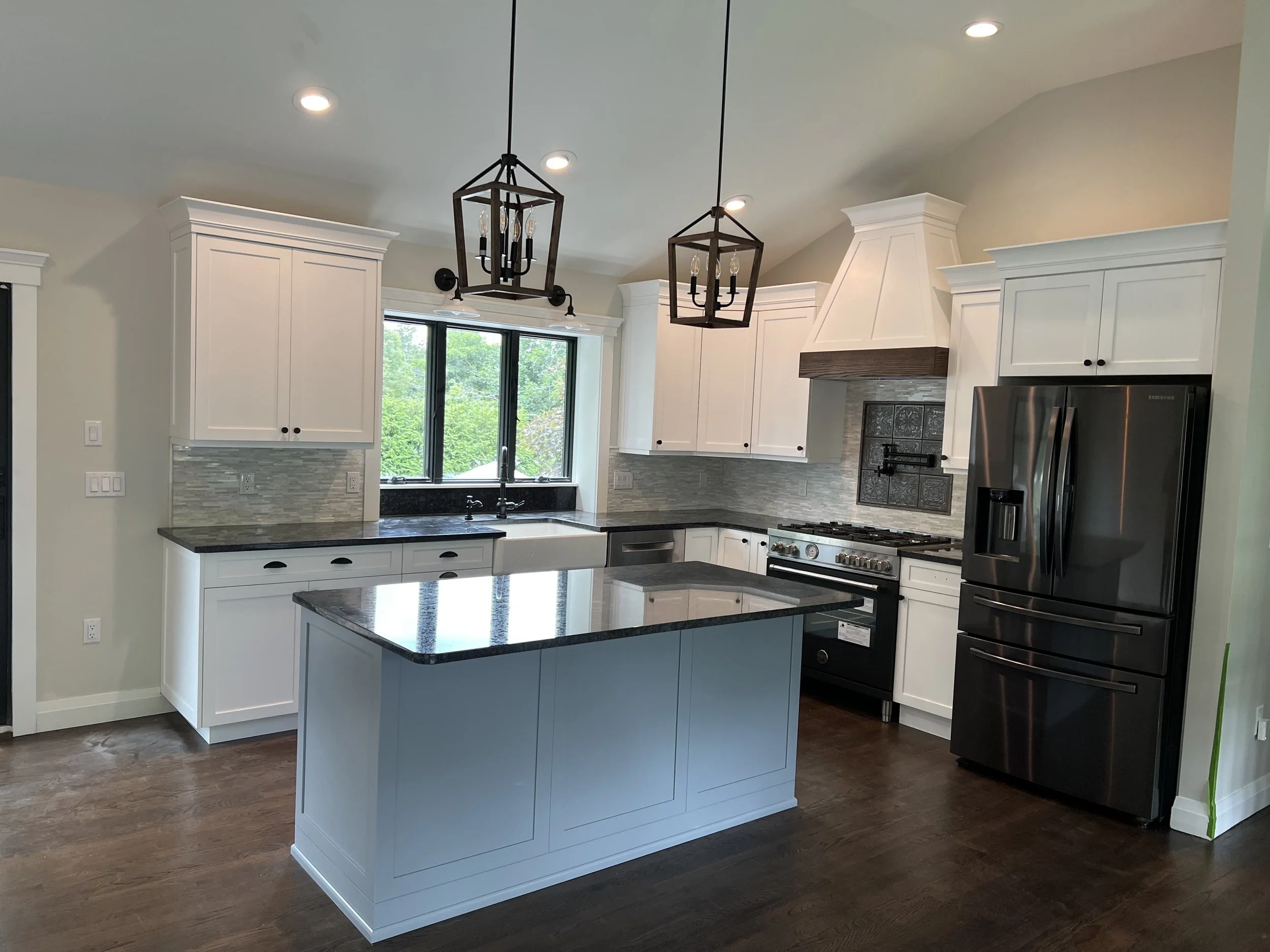 Modern kitchen with white cabinets, black countertops, a black refrigerator, stainless steel appliances, dark wood flooring, and two black pendant light fixtures over an island.