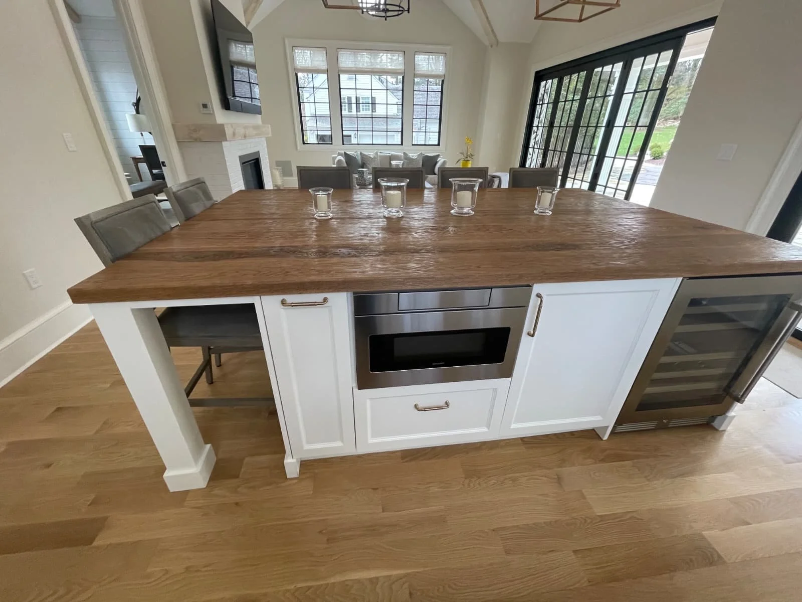 A modern kitchen island with a wooden countertop, white cabinet base, and built-in microwave. The island has barstools on one side and a wine fridge on the other. In the background, a dining and living area with large windows and a sliding glass door
