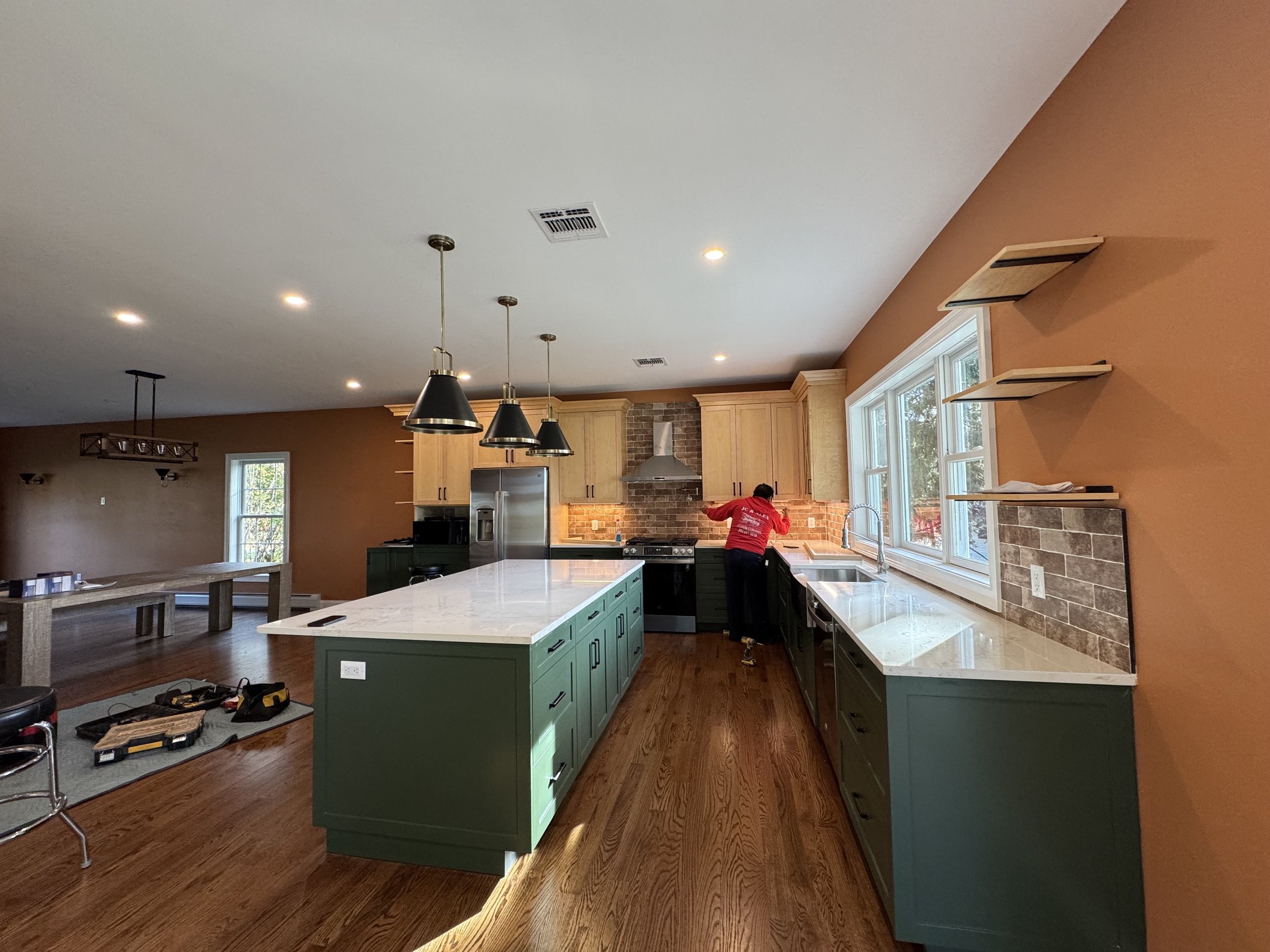 Modern kitchen under construction with green cabinets, white marble island, brick backsplash, and a person working near the sink.