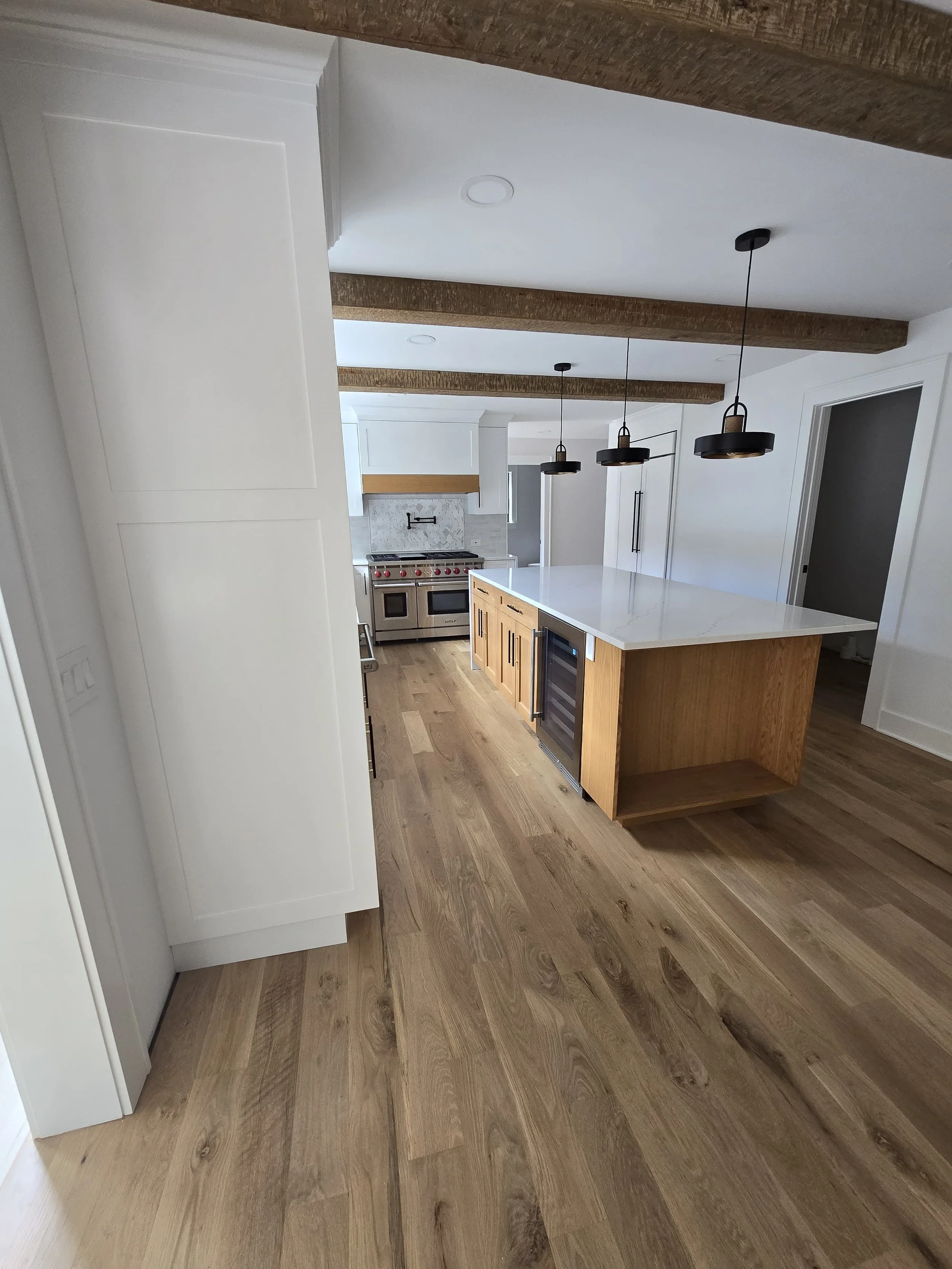 New modern kitchen featuring a large white marble island with wooden cabinet base, three black pendant lights hanging above, hardwood floors, white cabinetry, and a stainless steel stove in the background.