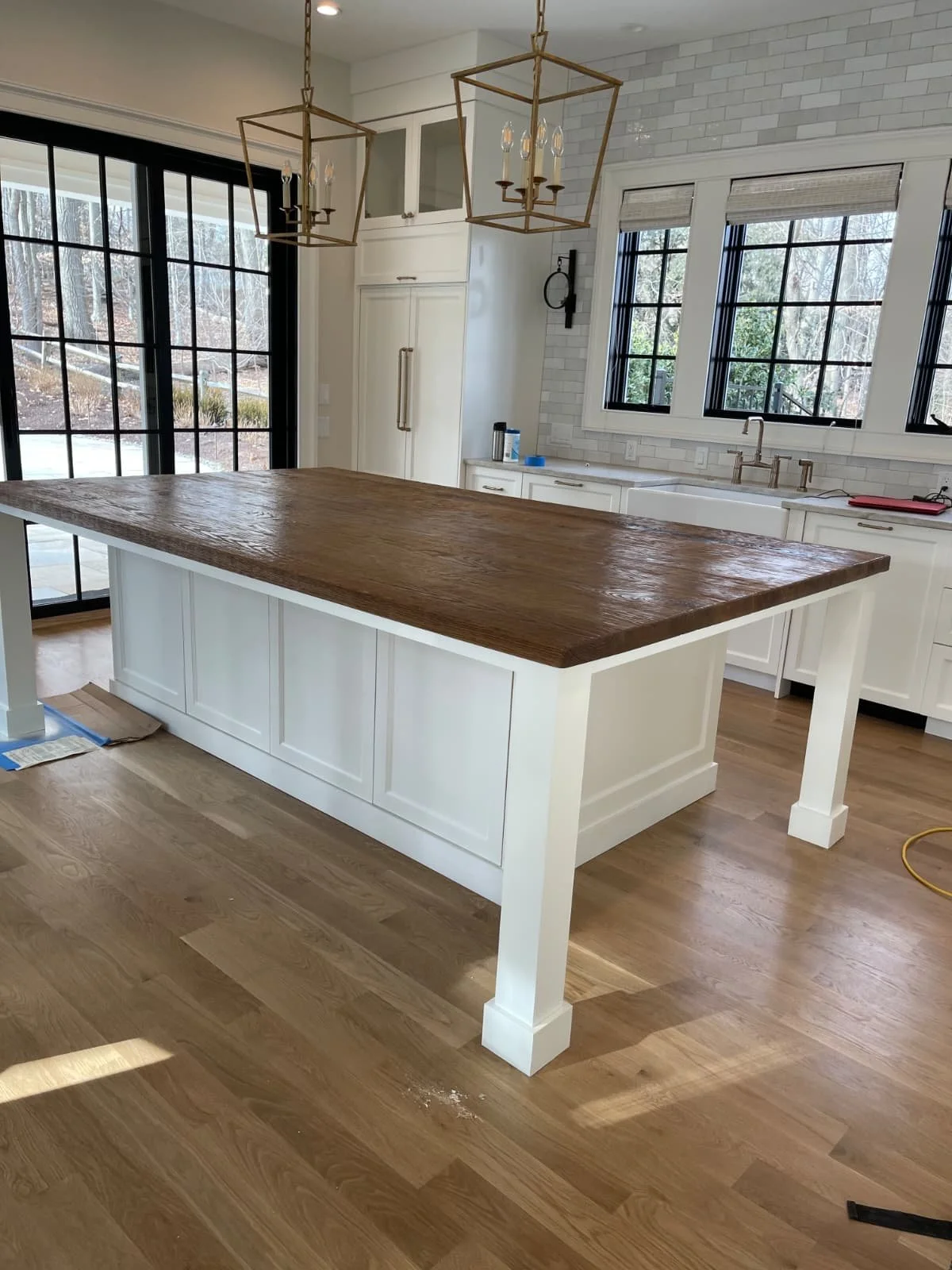 Interior view of a kitchen with a central island with a dark wooden countertop and white base. Behind it are white cabinets, a sink with a cabinet, and large windows. Two gold-colored chandeliers hang from the ceiling.