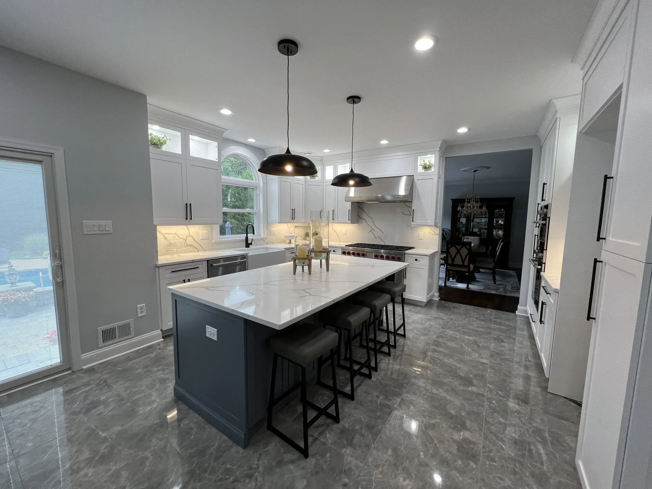 Modern kitchen with white cabinetry, marble backsplash, grey island with black barstools, black pendant lights, grey marble floor, and a view into a dining room.